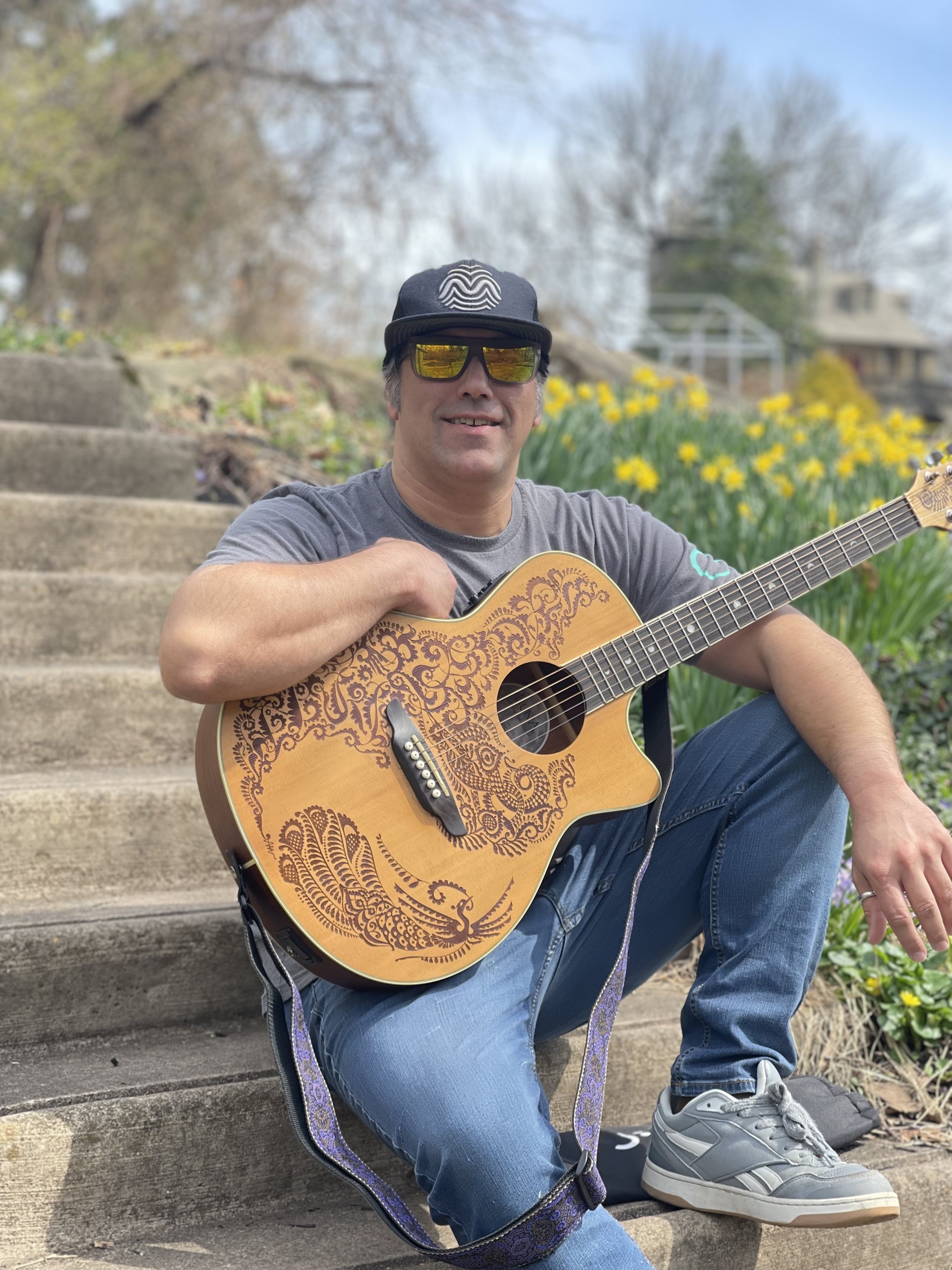 A man sitting on outdoor steps playing an acoustic guitar, wearing sunglasses, a baseball cap, and casual clothing, with flowers and a house in the background.