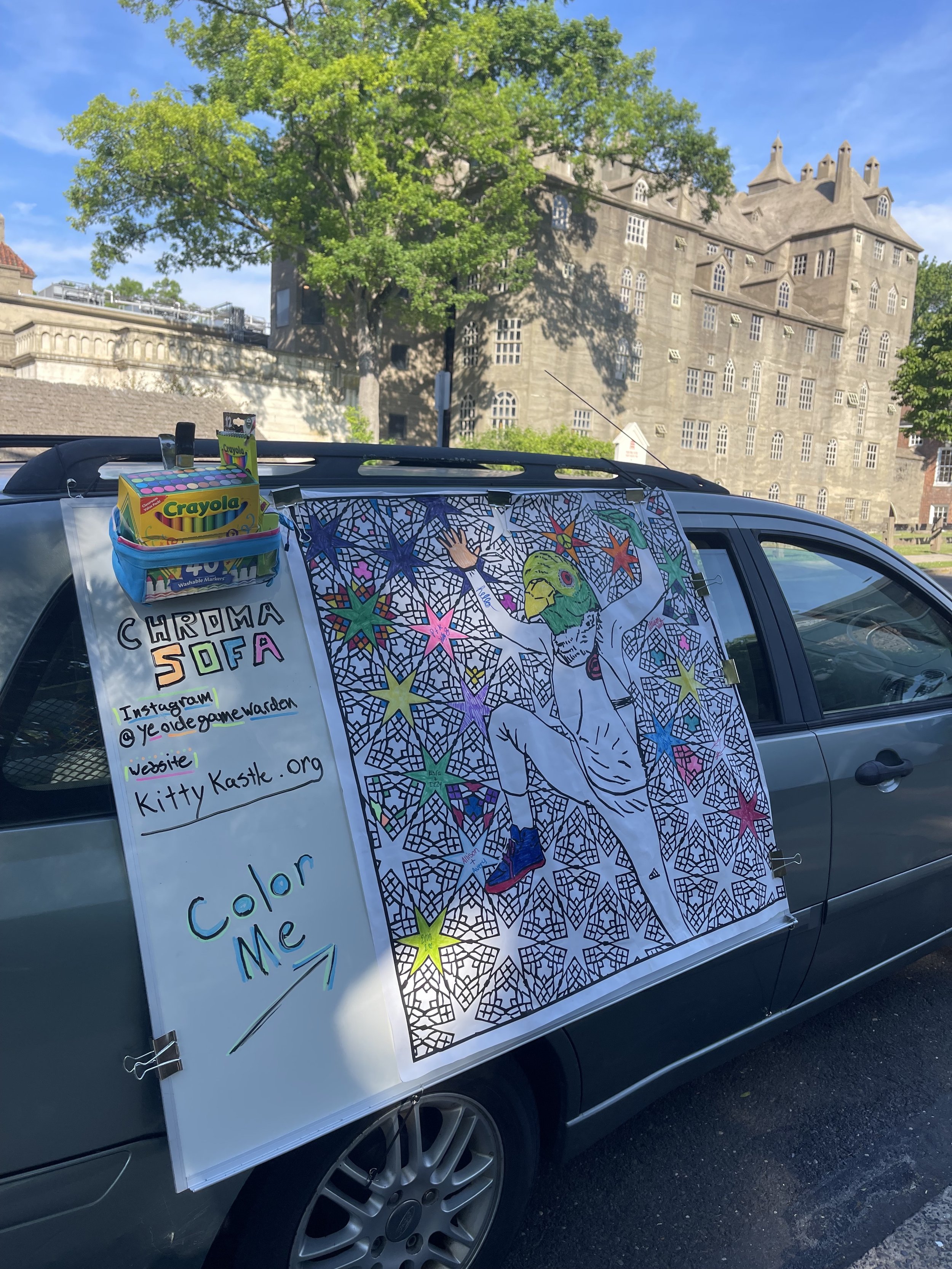 A sign on a car promoting a coloring activity for children. The sign features a large, colorful coloring page of a parrot surrounded by stars and other designs, with handwritten notes providing the Instagram handle @yeoldegamewarden and the website k
