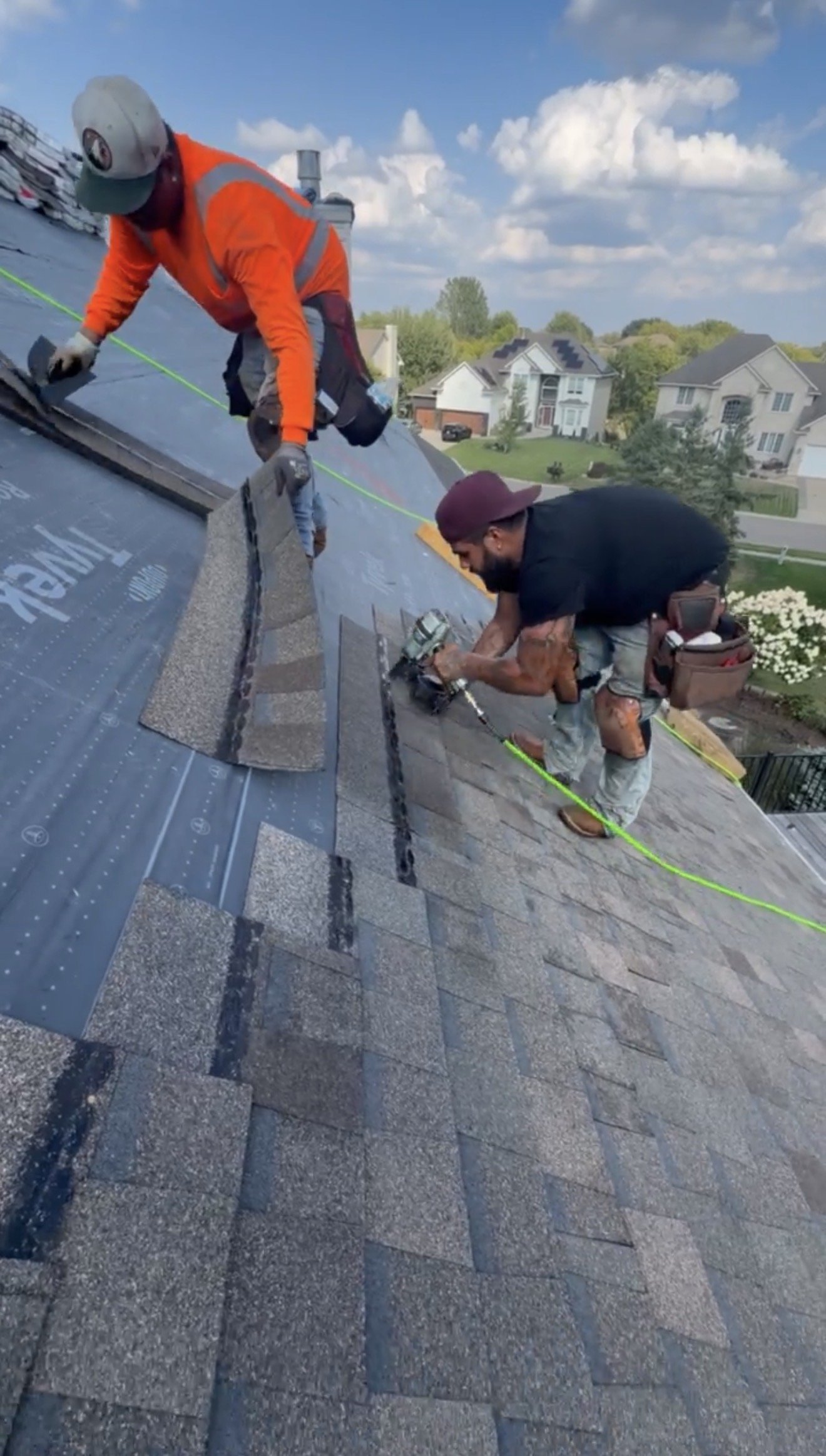 Two workers on a sloped rooftop installing shingles. One is kneeling with a power tool, the other is bending over, both wearing safety gear. In Maple Grove MN