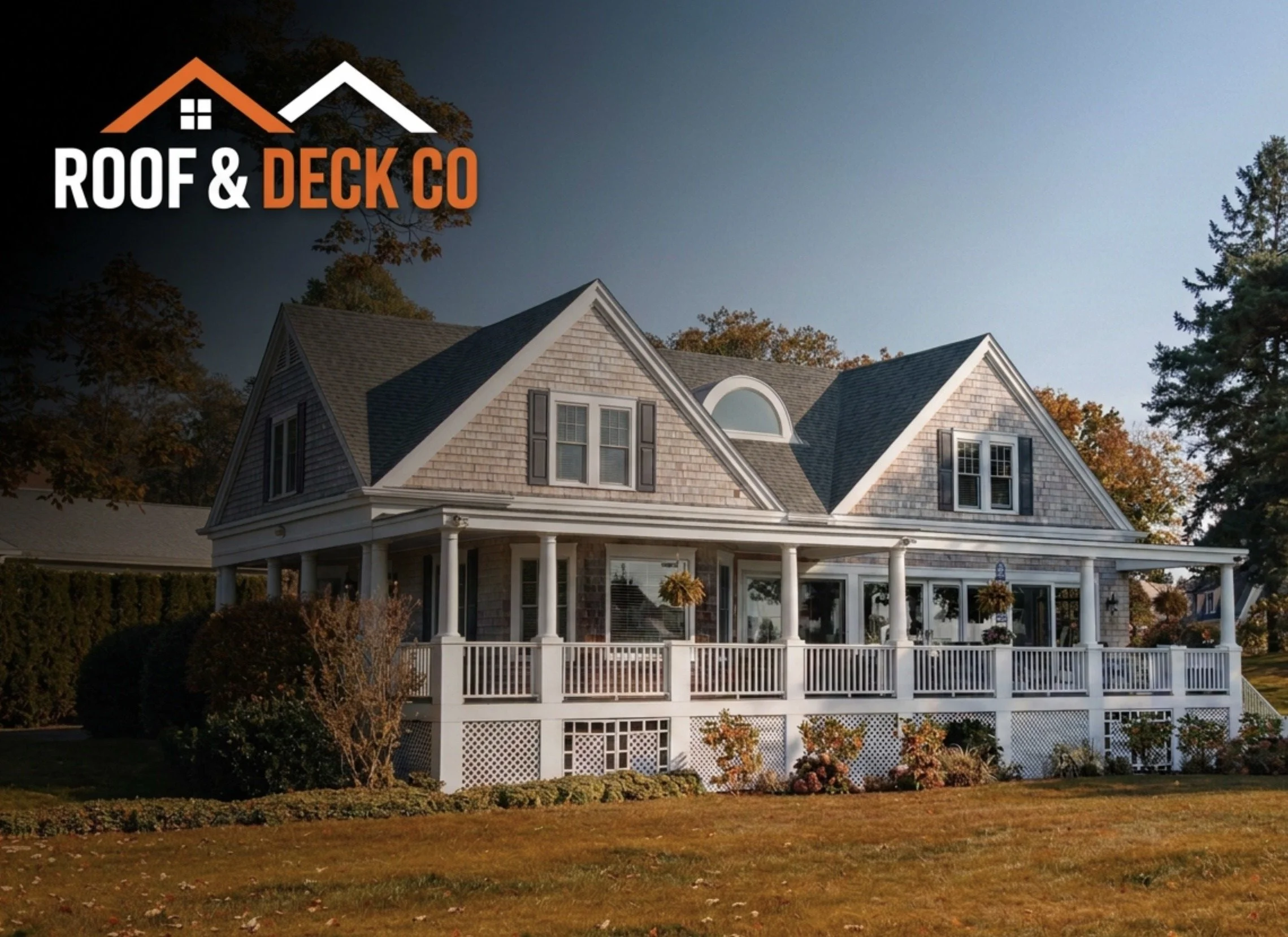 A large house with gray shingles, a porch with white columns, a white picket fence, and surrounding trees with fall foliage during daytime.