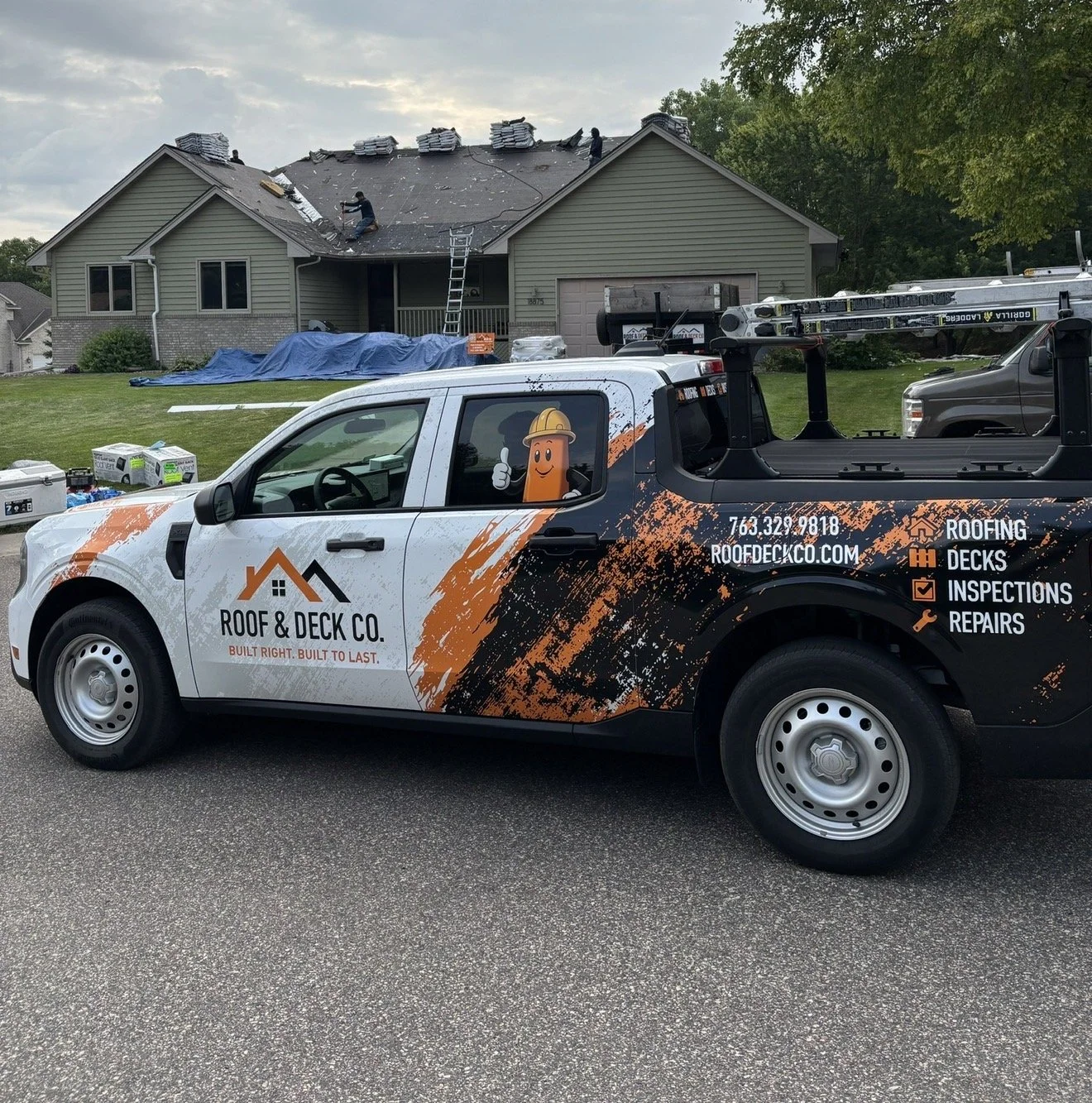 A pickup truck from Roof & Deck Co. parked in front of a house, with contractors working on the roof. Based in Maple grove MN