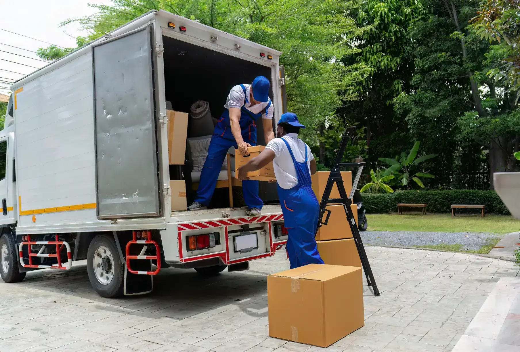 Two movers in blue uniforms loading cardboard boxes into a moving truck parked on a driveway surrounded by greenery.