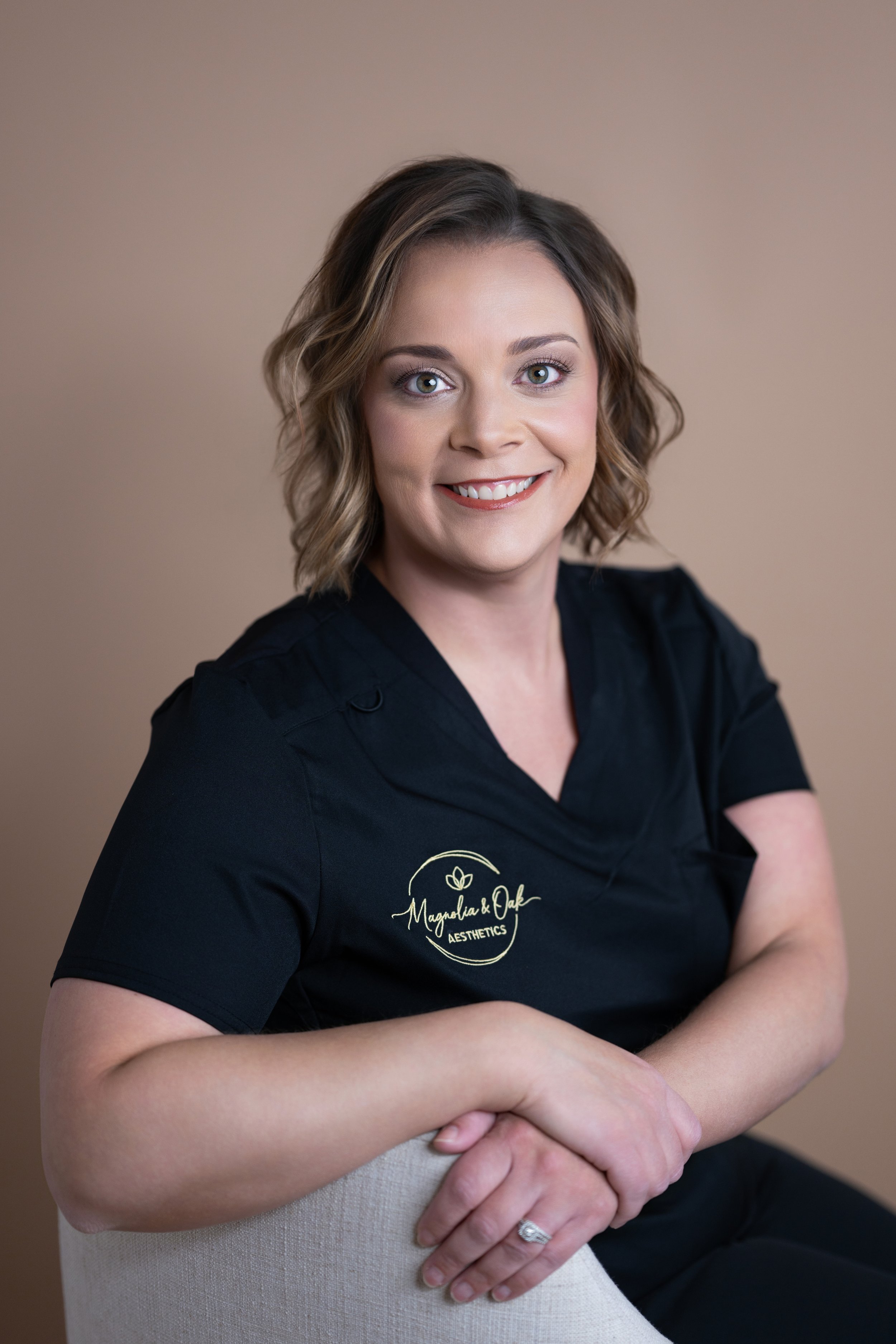 Portrait of a woman with shoulder-length wavy brown hair, wearing a black shirt with a logo that reads 'Magnolia & Oak Aesthetics,' sitting on a beige chair with a warm neutral background.