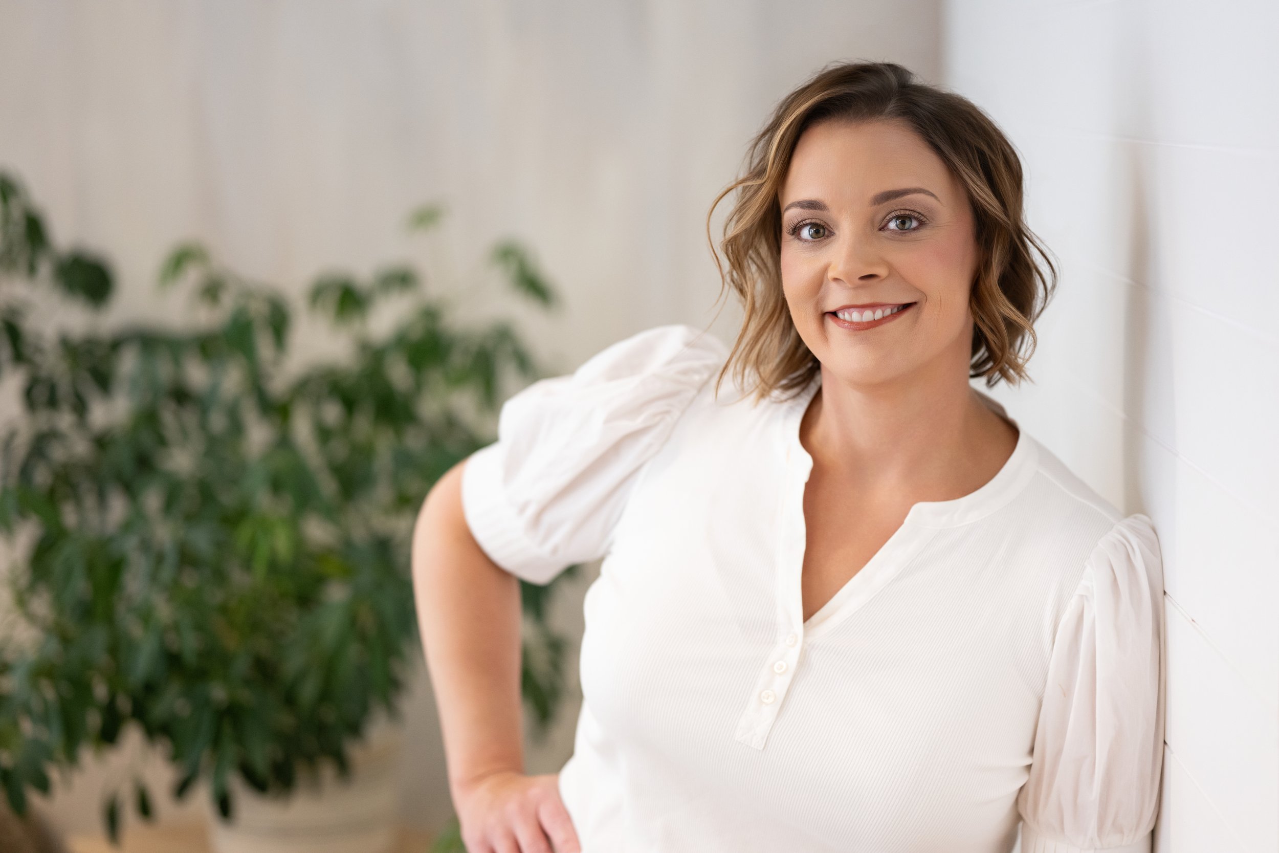 Woman with short brown hair smiling and wearing a white top standing in front of a white wall and green plant.