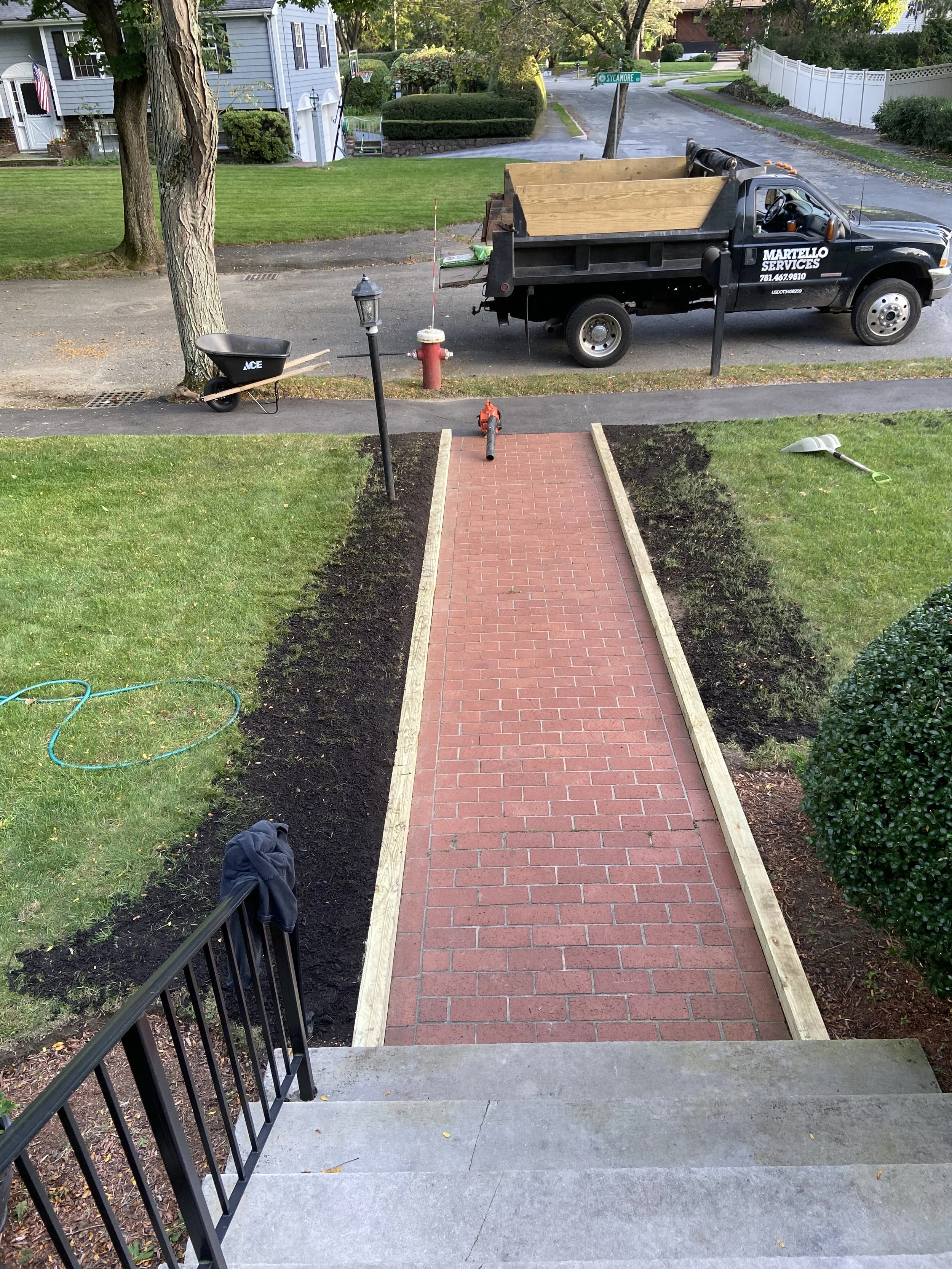 View from the top of outdoor concrete stairs leading down to a sidewalk and a newly paved red brick pathway, with a black metal fence on the left and a garden with dark soil on both sides. In the background, there is a neighborhood street with a parked work truck labeled "Martello Services," a wheelbarrow, a garden shovel, a fire hydrant, and a street sign indicating the intersection of Sycamore and another street, with trees and houses lining the street.