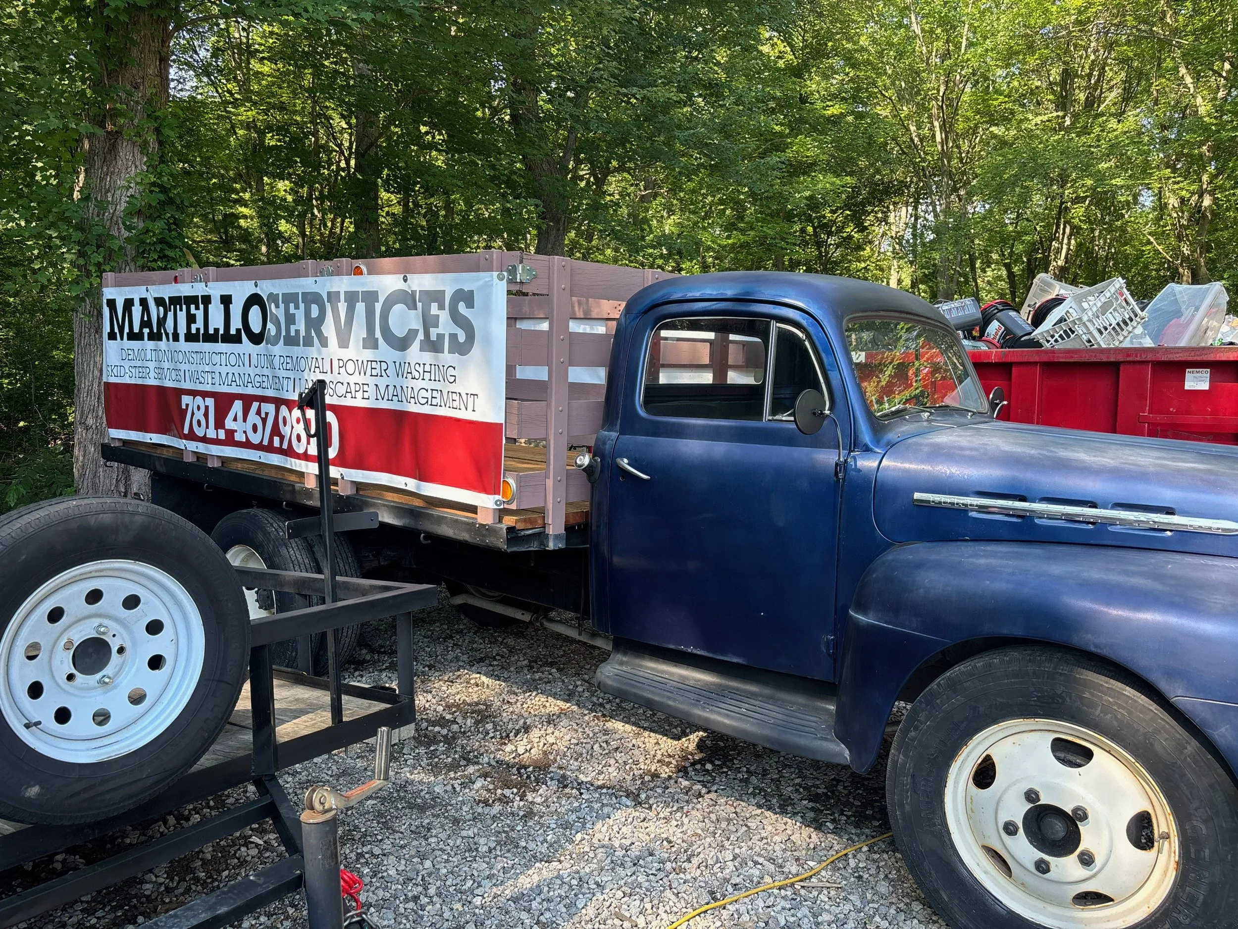 An old blue pickup truck with a flatbed, parked outdoors among trees, carrying a red container filled with various tools and equipment, and a sign on the side for Martello Services.