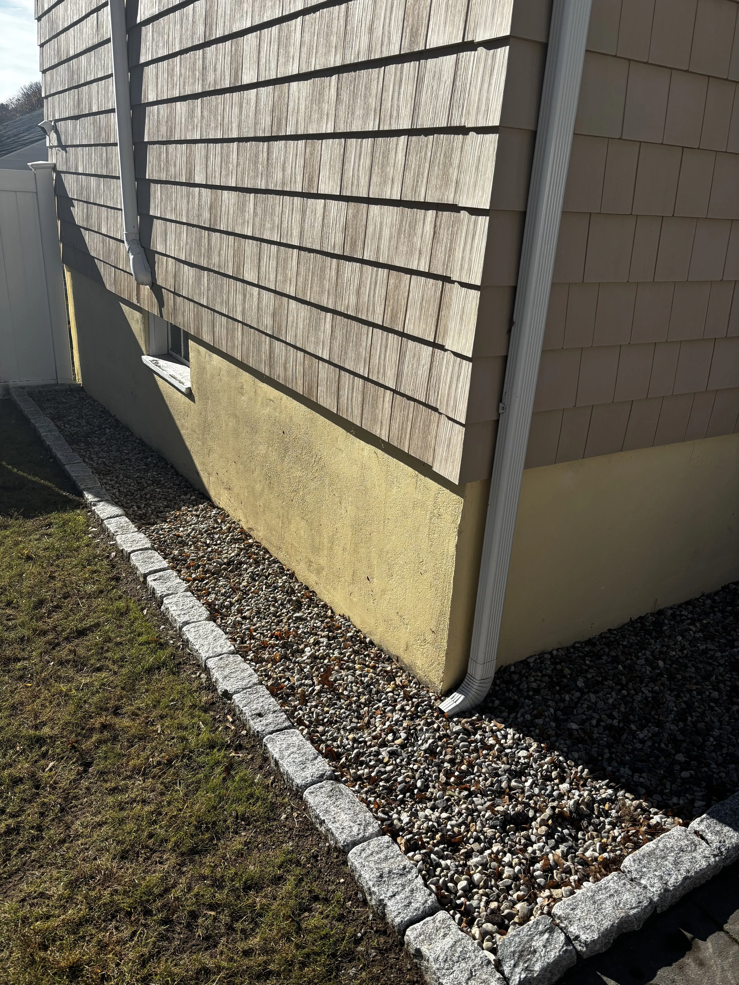 Side view of a house with wooden and vinyl siding, a downspout, a small window with a concrete sill, and a gravel flower bed edged with large rocks, with a grassy area in the foreground.