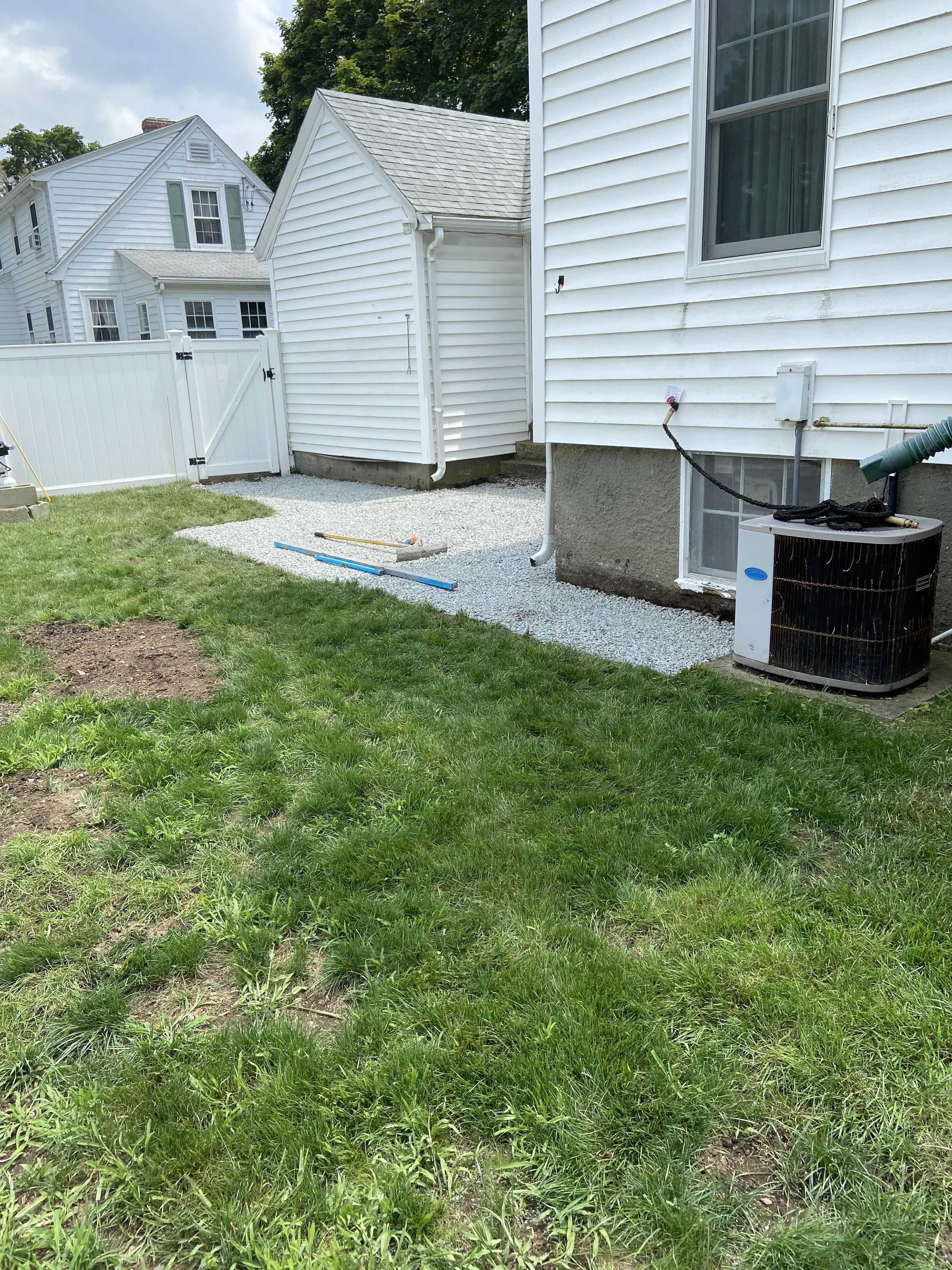 Backyard with green grass, a white house with siding, a small gravel area, a white fence and a few tools and equipment including a blue level, a black and white pipe, and a large outdoor air conditioning unit.