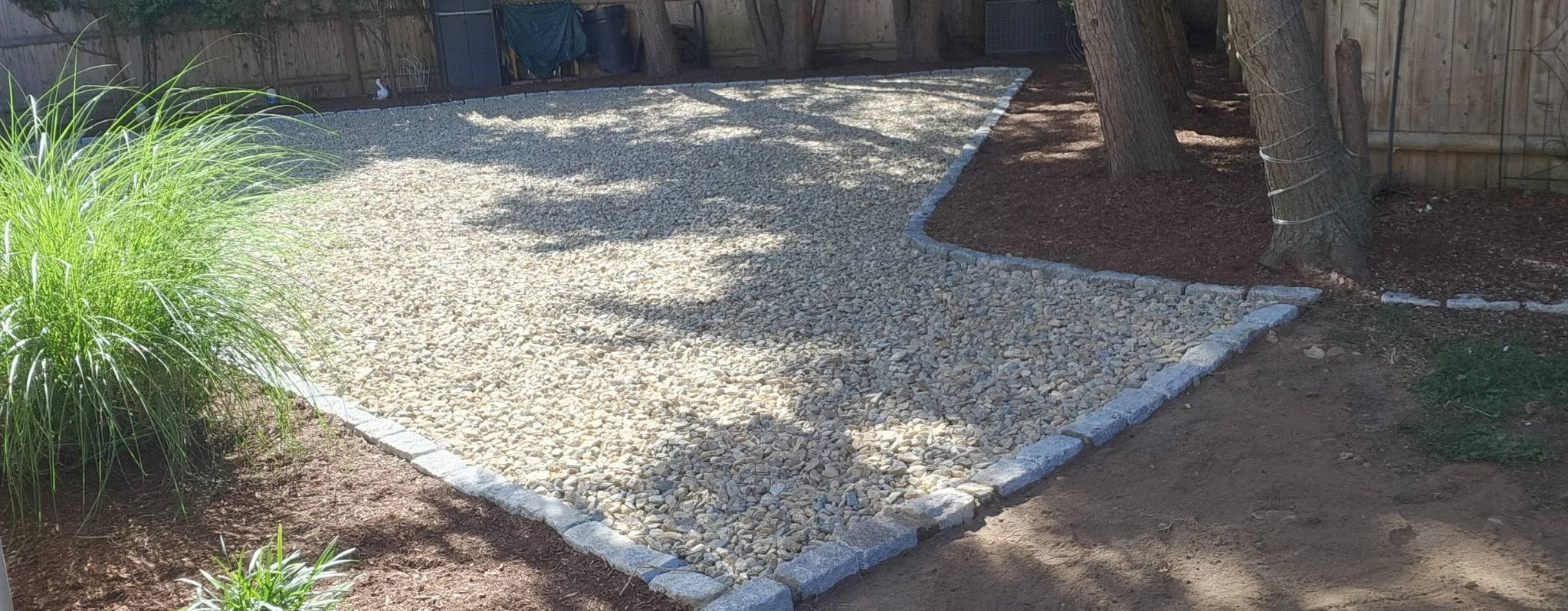 Backyard with a landscaped area featuring a gravel bed bordered by stone bricks, a large tree with ties around its trunk, a wooden fence, and various plants and trees.