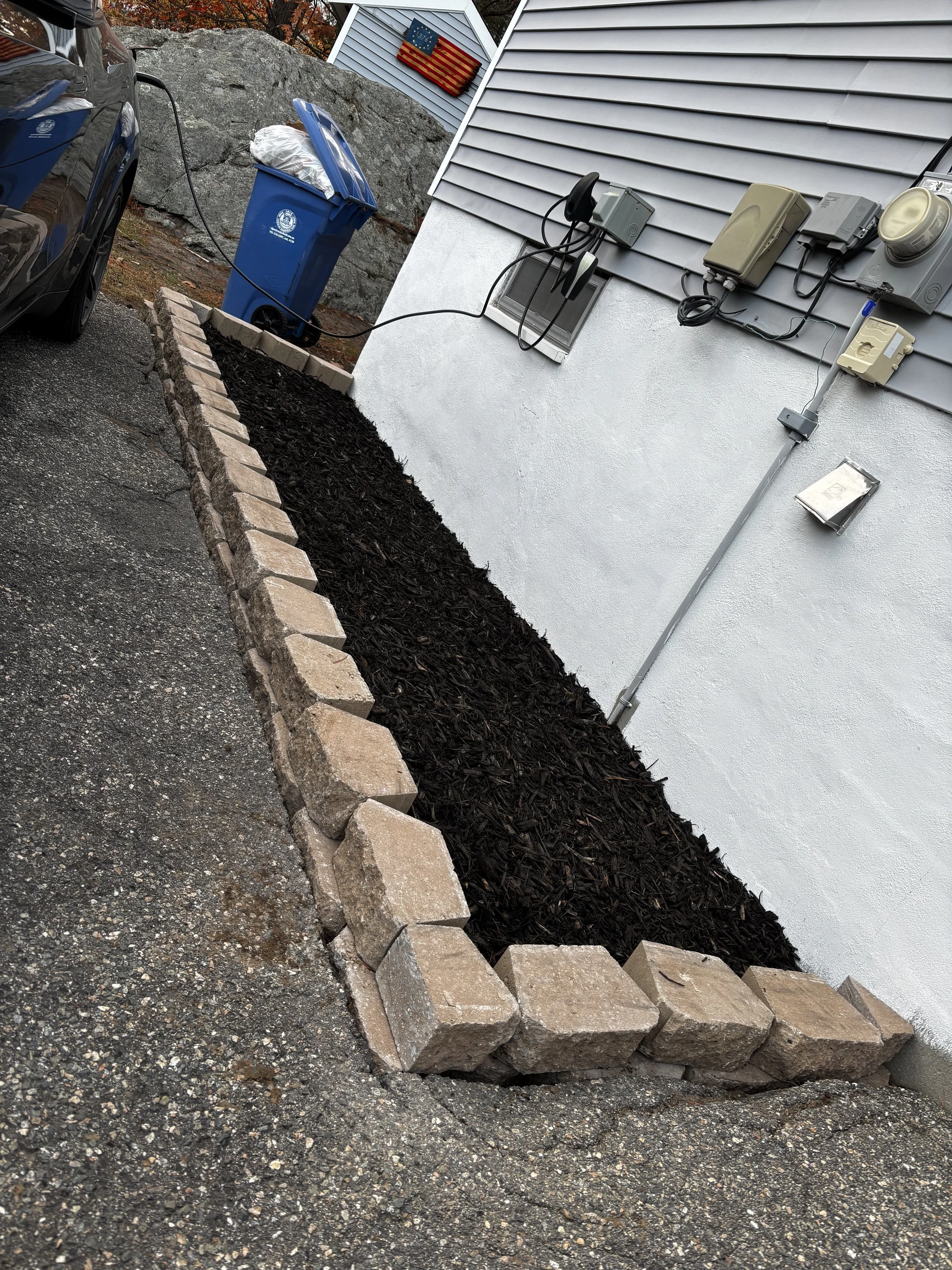 A parking area with a black car plugged into an exterior electrical outlet, a blue recycling bin filled with garbage, a small flower bed with black mulch bordered by stone bricks, and the side of a house with electrical meters and wiring, American flags, and a window.
