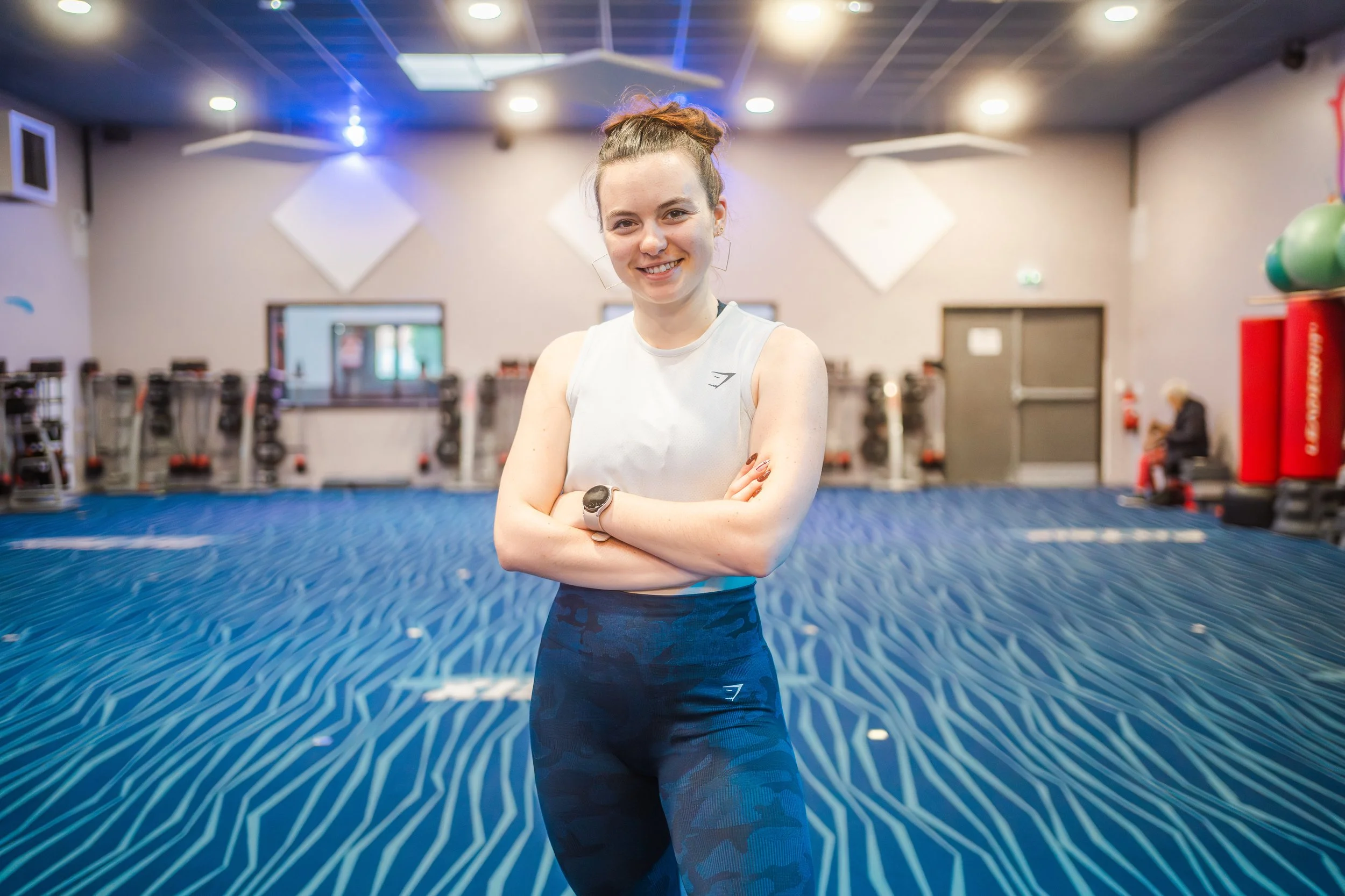 Une femme souriante dans une salle de sport avec un sol bleu, portant un débardeur blanc et un pantalon de sport bleu. Elle a les bras croisés.