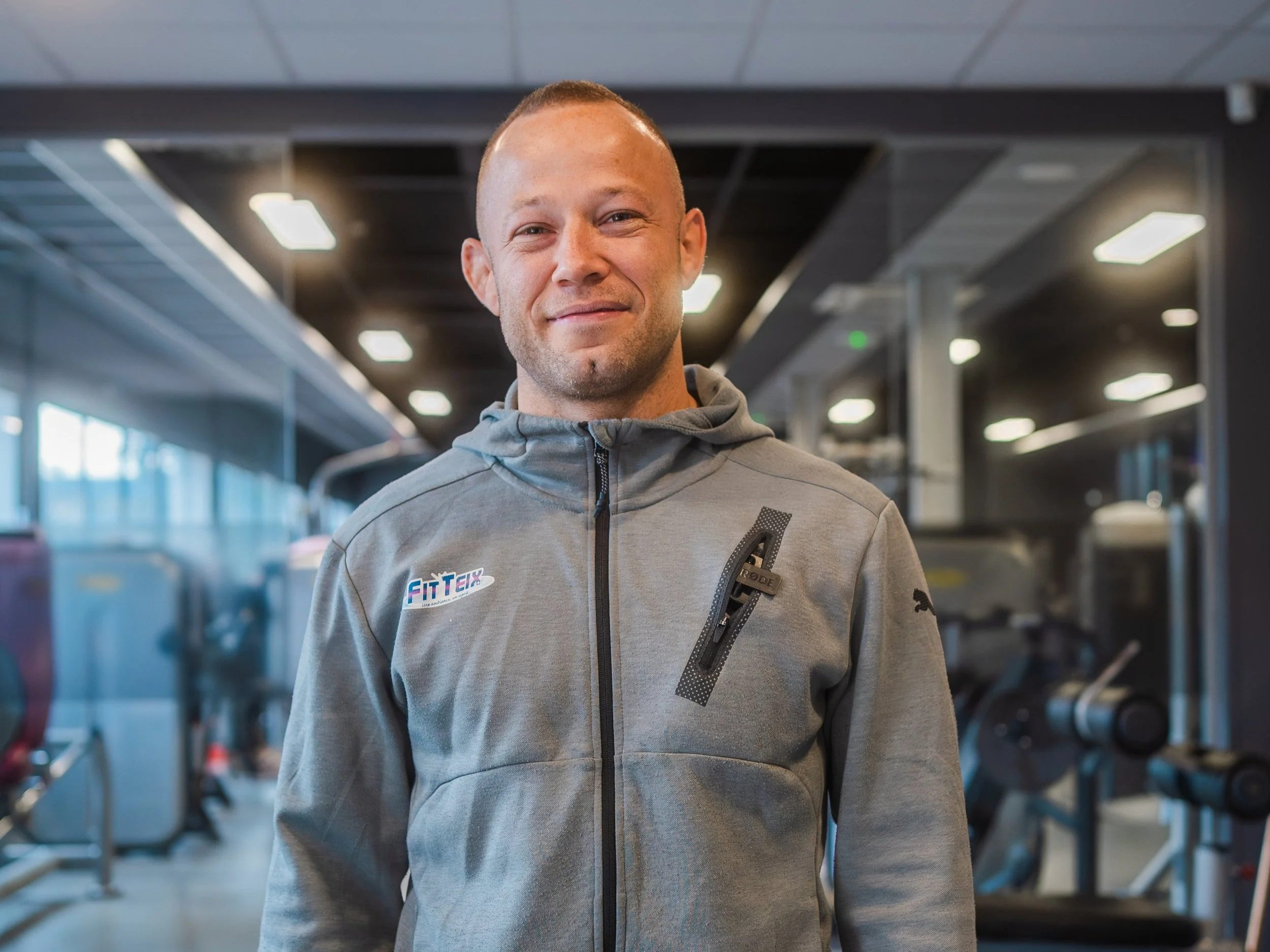 Un homme dans une salle de sport, portant un sweat à capuche gris avec un logo "FitTek". Il sourit, et l'arrière-plan montre des équipements de gym et des lumières au plafond.