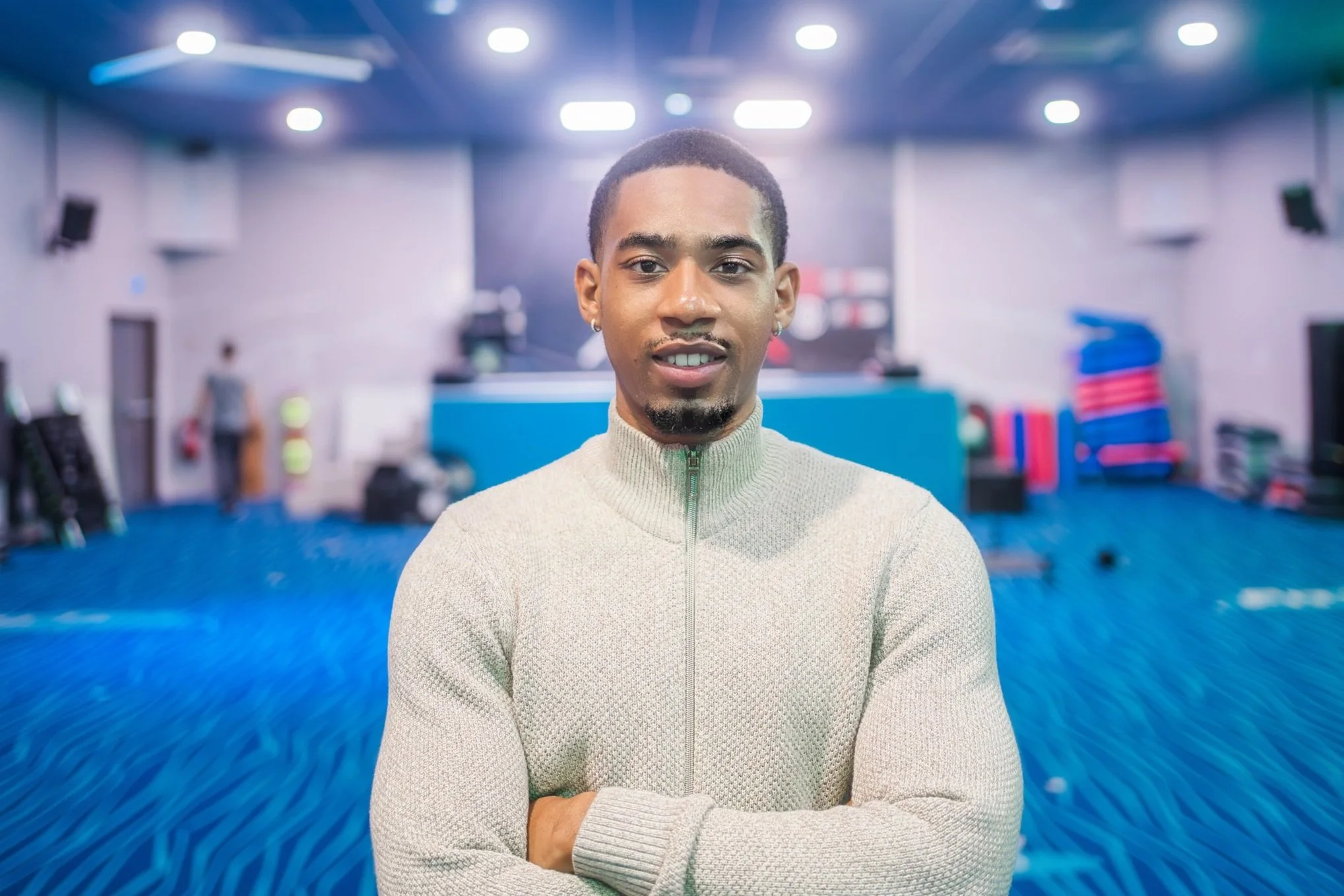 Un homme souriant avec un pull beige, debout dans une salle avec un sol bleu et des équipements sportifs en arrière-plan flou.