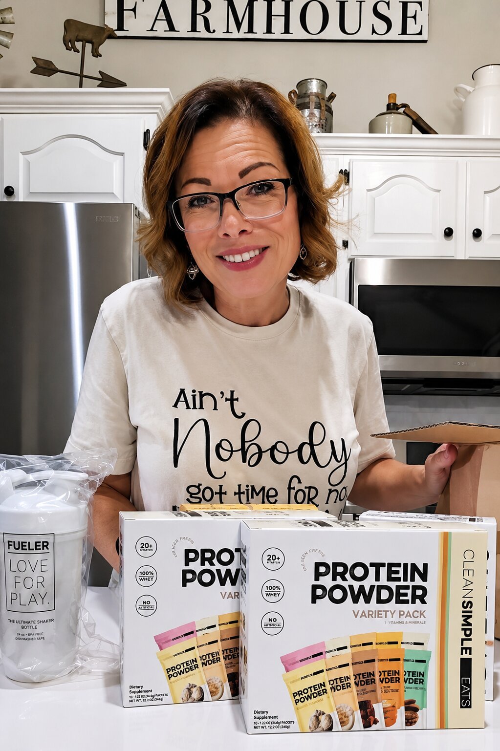 A woman with glasses and short brown hair smiling in a kitchen, wearing a white T-shirt that says 'Ain't Nobody got time for no'. She is standing behind a table with boxes of protein powder, a shaker bottle, and some packets of protein powder flavor samples.