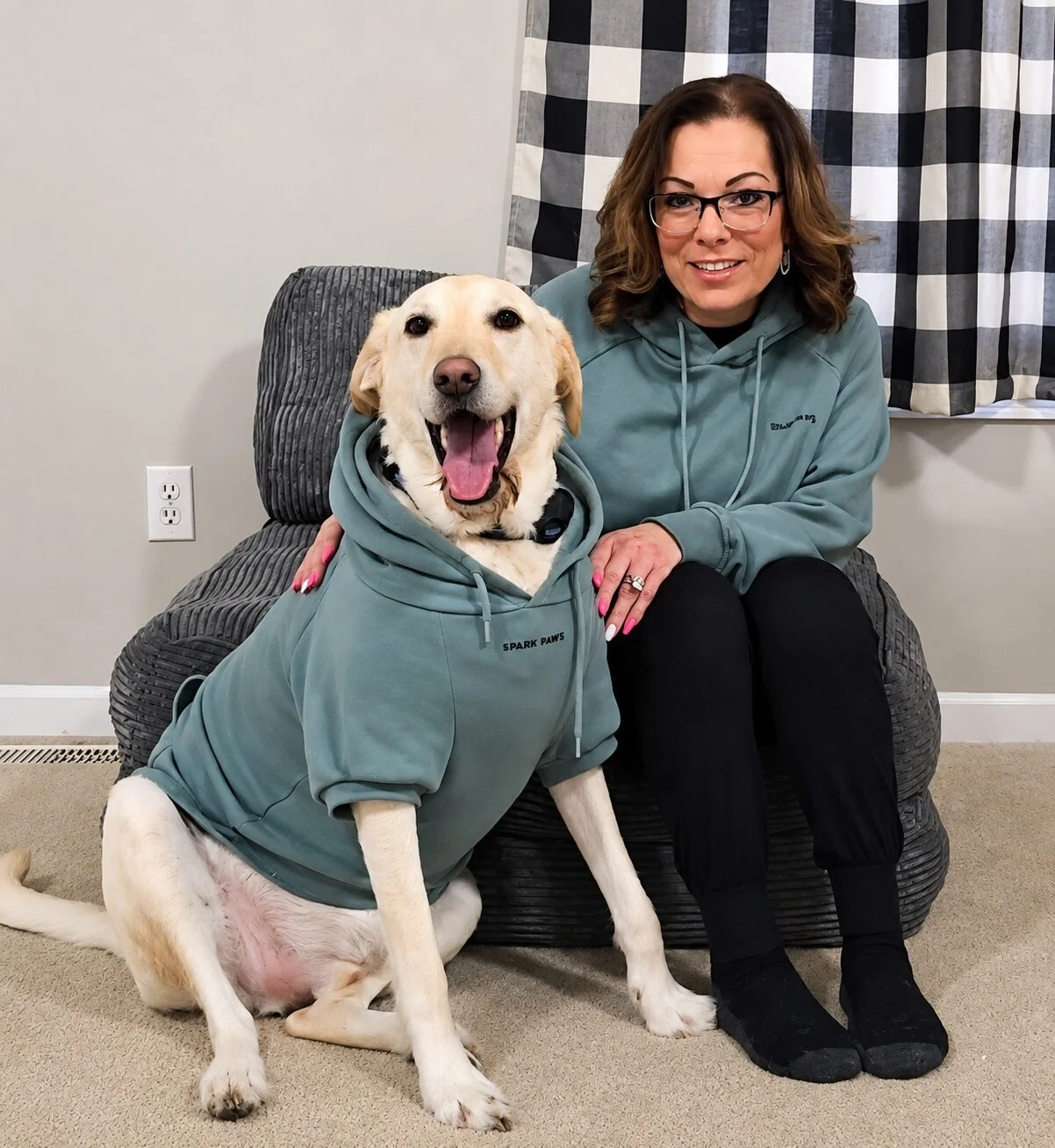 A woman with glasses sitting on a gray armchair beside a happy Labrador retriever dog wearing a matching hoodie. The woman has brown hair, is smiling, and is dressed in a teal hoodie and black pants. The dog, sitting on the floor, has its mouth open, showing its tongue, and is also wearing a teal hoodie. The background features a black and white checkered curtain, and an electrical outlet is visible on the wall.