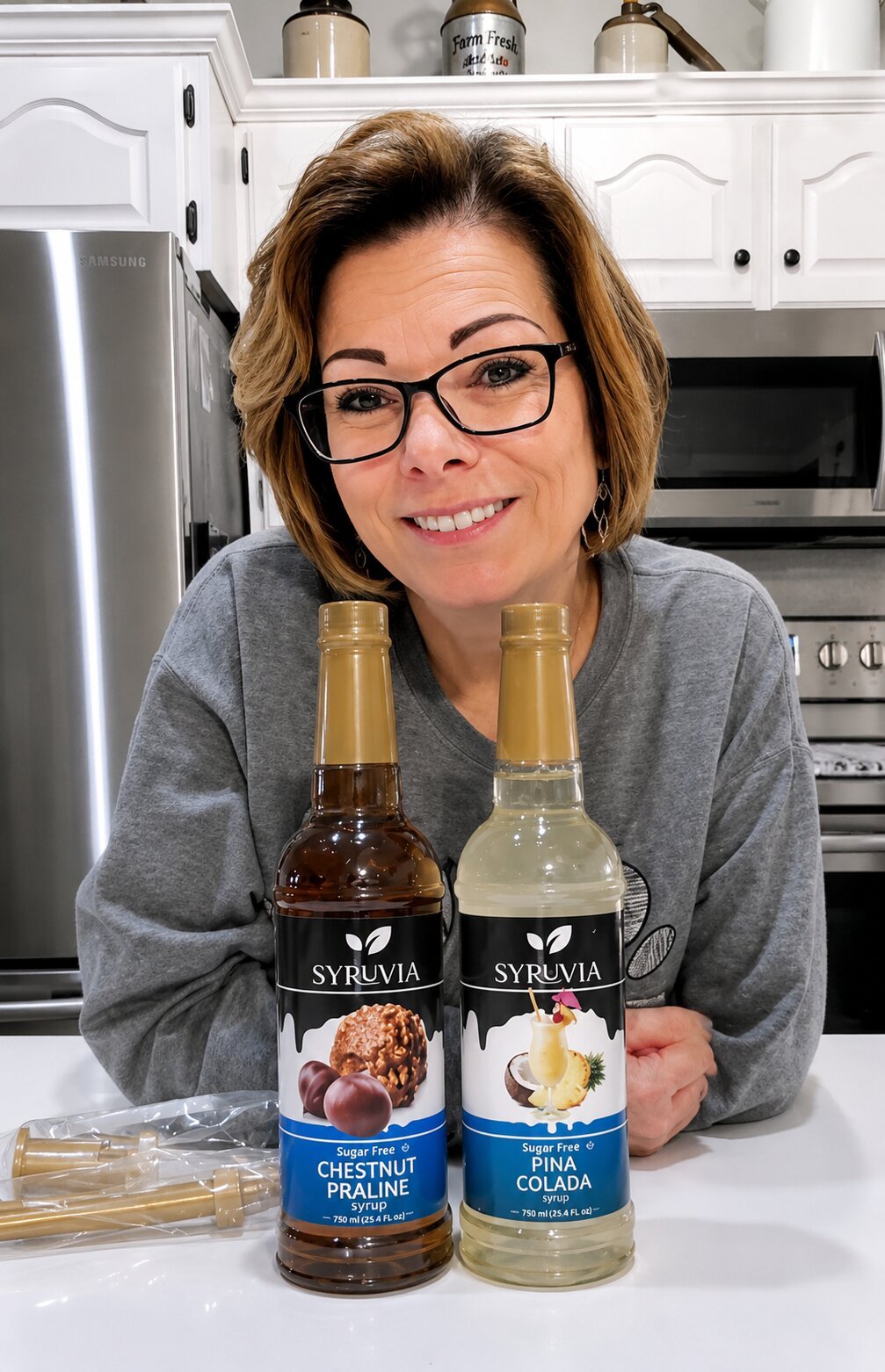 Smiling woman with glasses in a kitchen, displaying two bottles of syrup labeled 'Syruvia,' one Chestnut Praline flavor and one Pina Colada flavor.