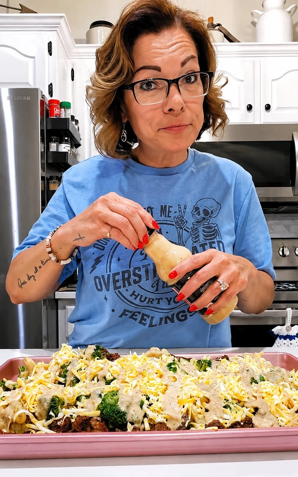 A woman with glasses and red nail polish is pouring sauce onto a tray of pasta with broccoli and shredded cheese in a kitchen.