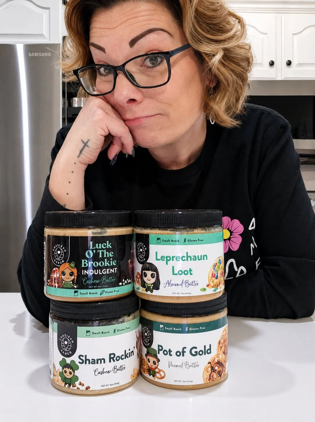 A woman with glasses, short wavy blonde hair, and a tattoo on her wrist is sitting at a table with four jars of flavored butters in front of her. The jars are labeled 'Luck O' The Brooke', 'Leprechaun Loot', 'Sham Rockin'', and 'Pot of Gold'. She is resting her chin on her hand and looking at the camera.