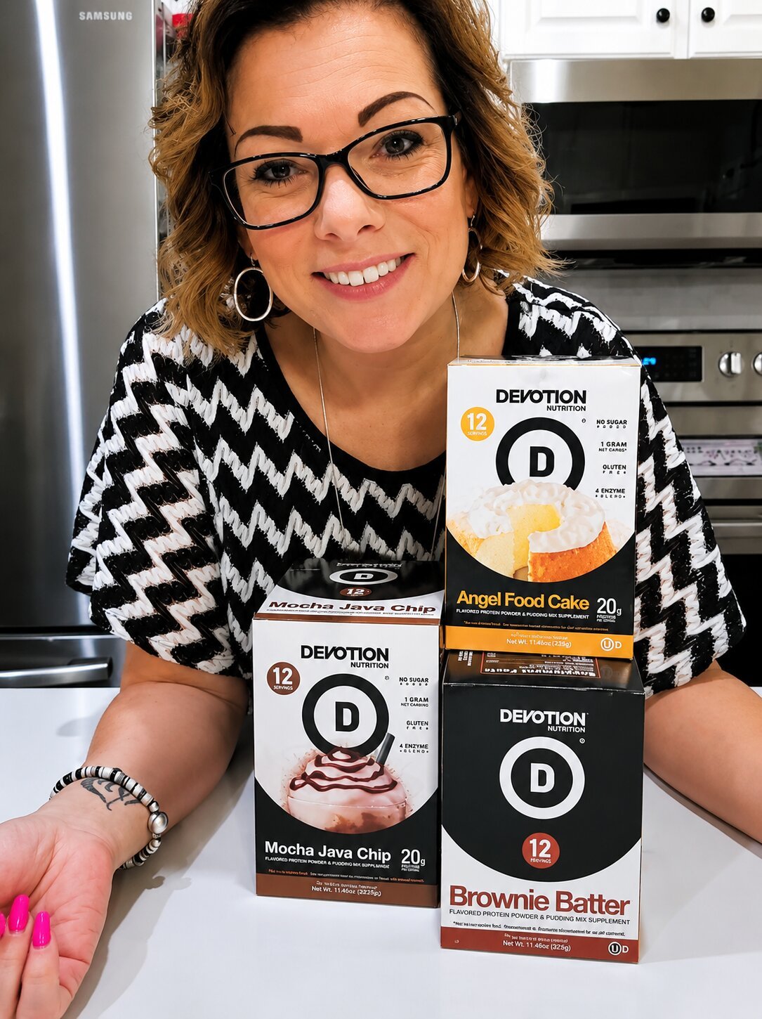 Woman with glasses smiling at camera, surrounded by Devotion Nutrition protein products, including Angel Food Cake, Mocha Java Chip, and Brownie Batter flavored protein powders.