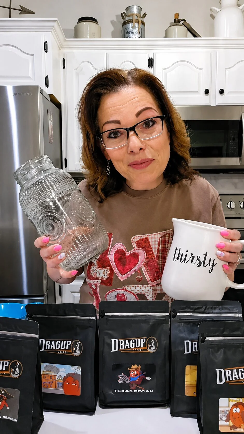 A woman with glasses holding a clear mason jar in one hand and a white mug with the word 'thirsty' in the other, standing in a kitchen with packaged coffee beans in front of her.