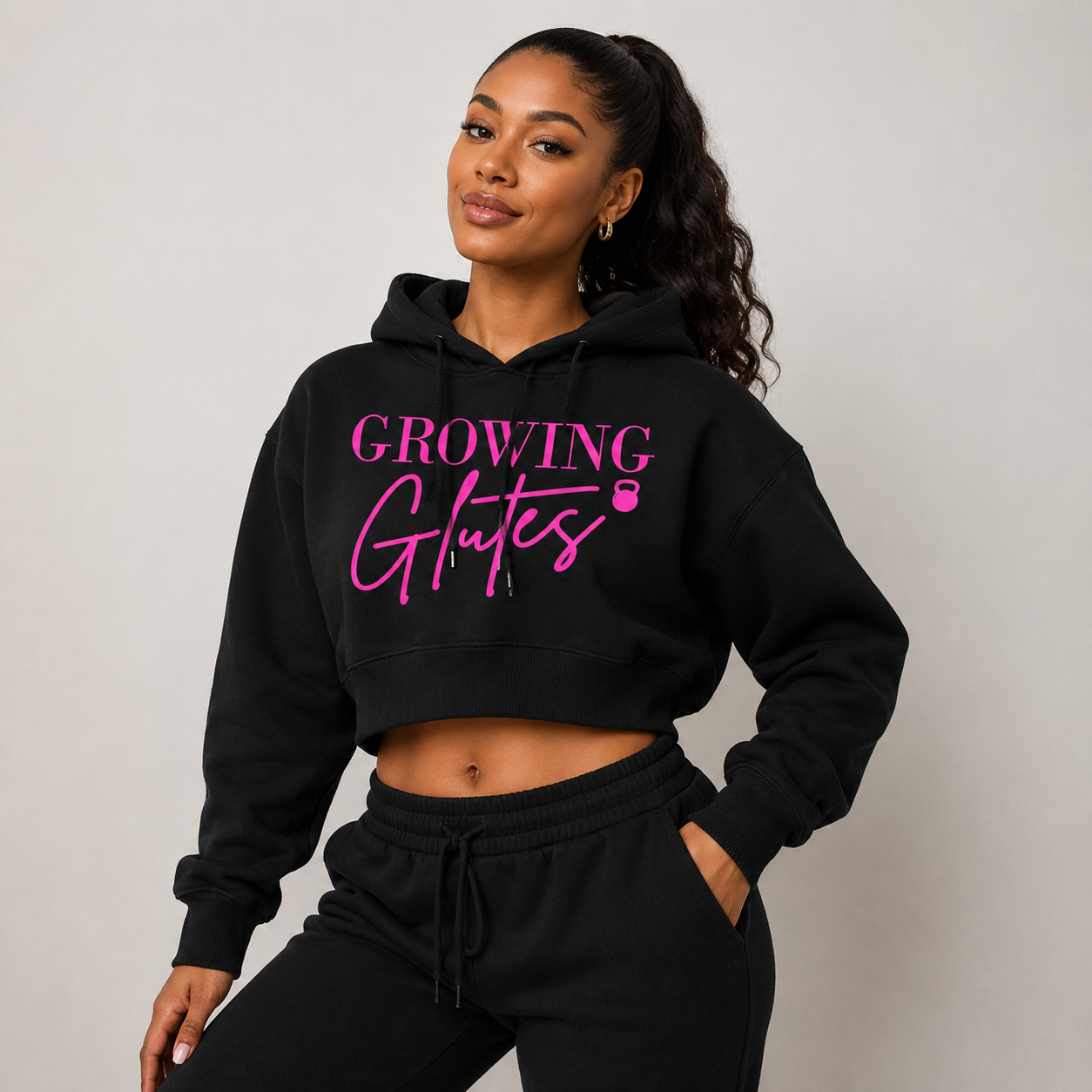 A woman wearing a black cropped hoodie with the text 'Growing Glutes' in pink, standing with one hand on her hip and smiling slightly.