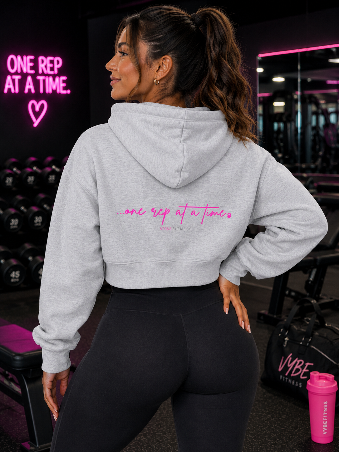 A woman with long brown hair tied in a ponytail, wearing a gray hoodie with pink text, stands in a gym. The hoodie says 'one rep at a time.' She is facing sideways, with one hand on her hip, and behind her is a neon sign that also says 'ONE REP AT A TIME' with a heart symbol. Gym equipment and a pink water bottle are visible in the background.
