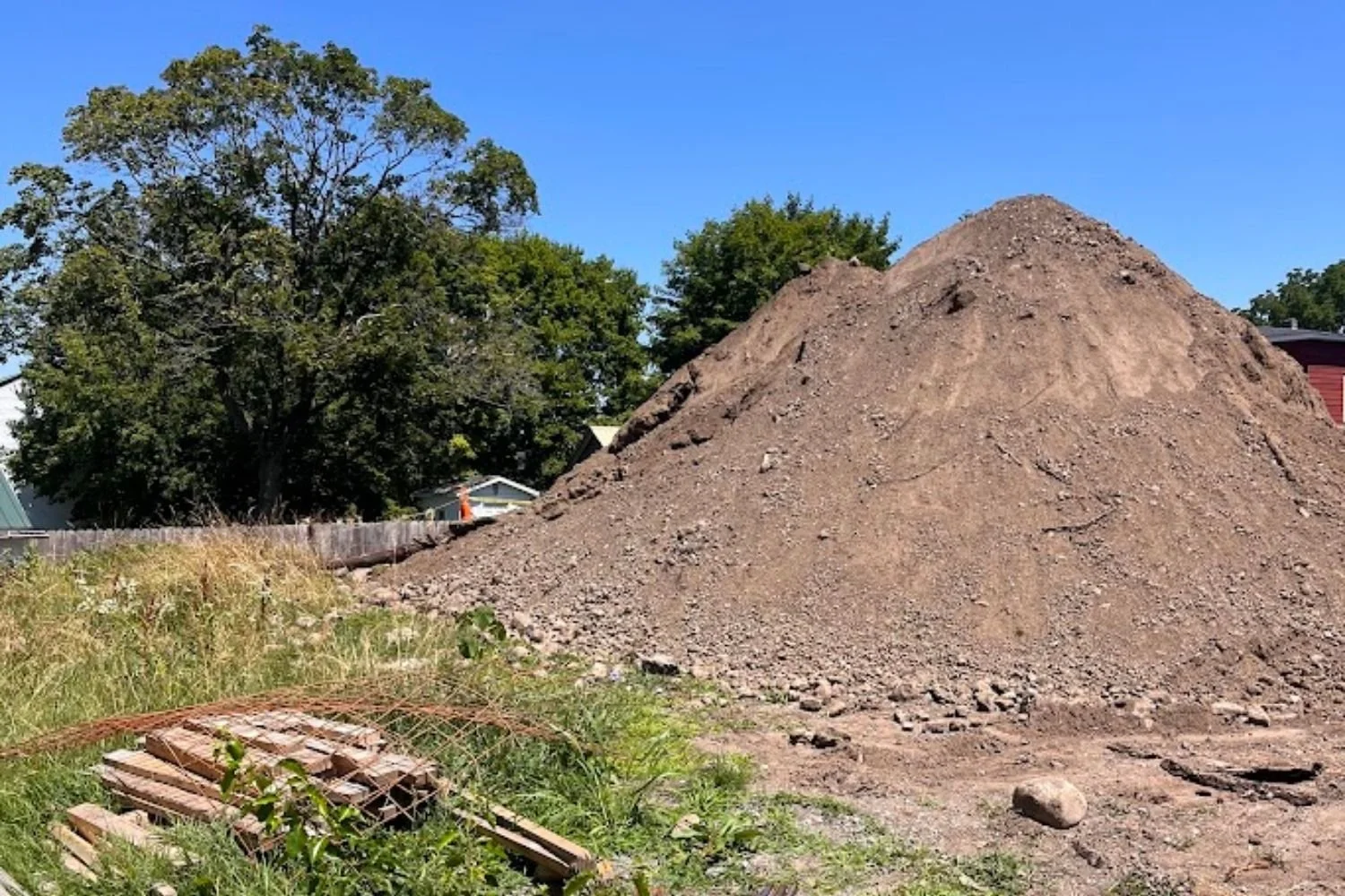 A large pile of dirt in a yard with trees and a house in the background on a sunny day.