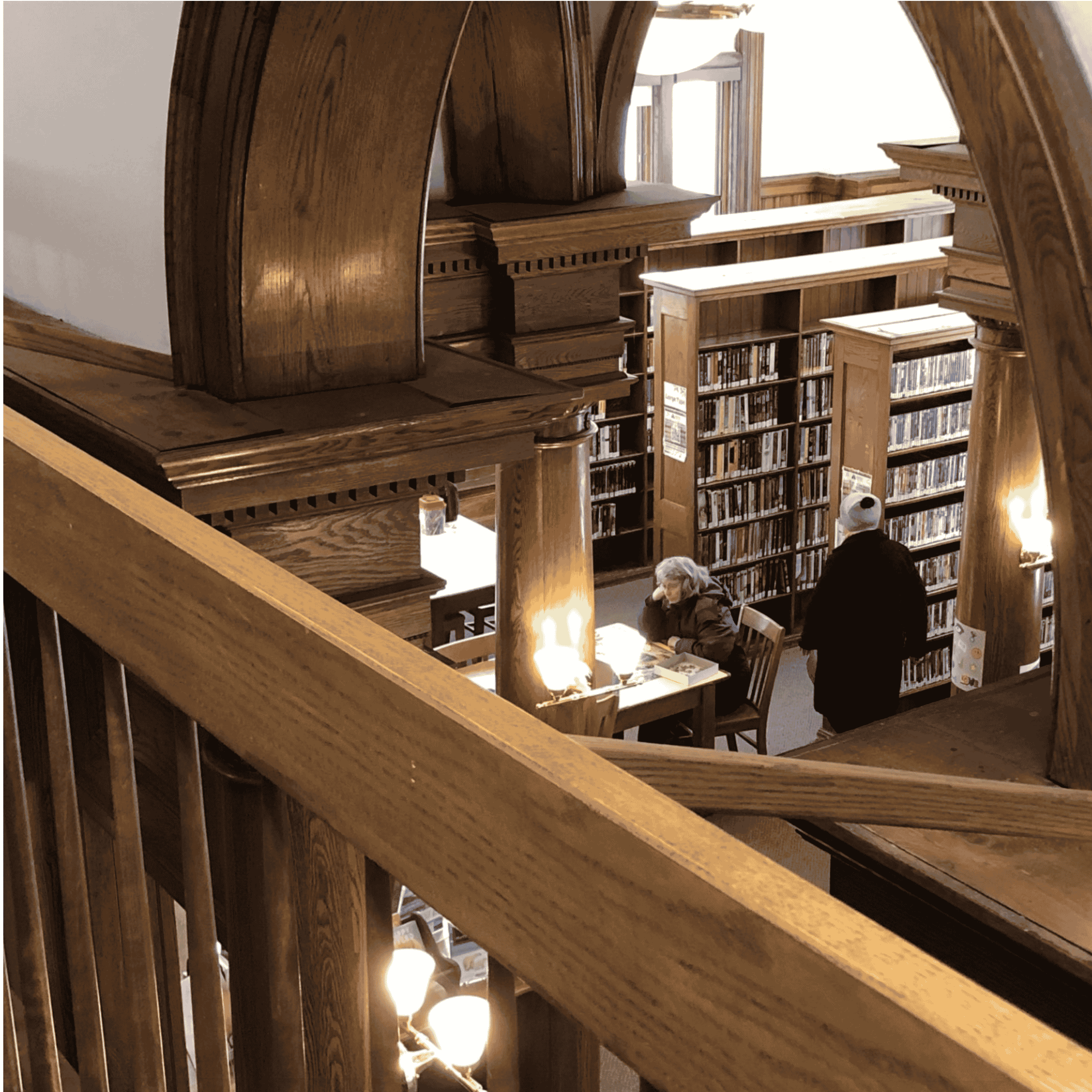 View of wooden stairs and balcony with a person sitting at a table below, wooden cabinets, and bookshelves filled with books in the background.