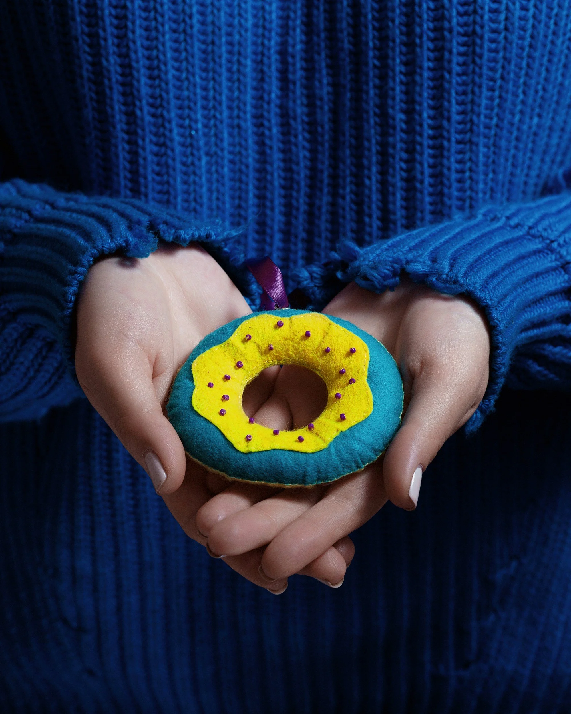 Close-up of a hand holding a felt donut with yellow icing and purple sprinkles, in front of a blue knit sweater.