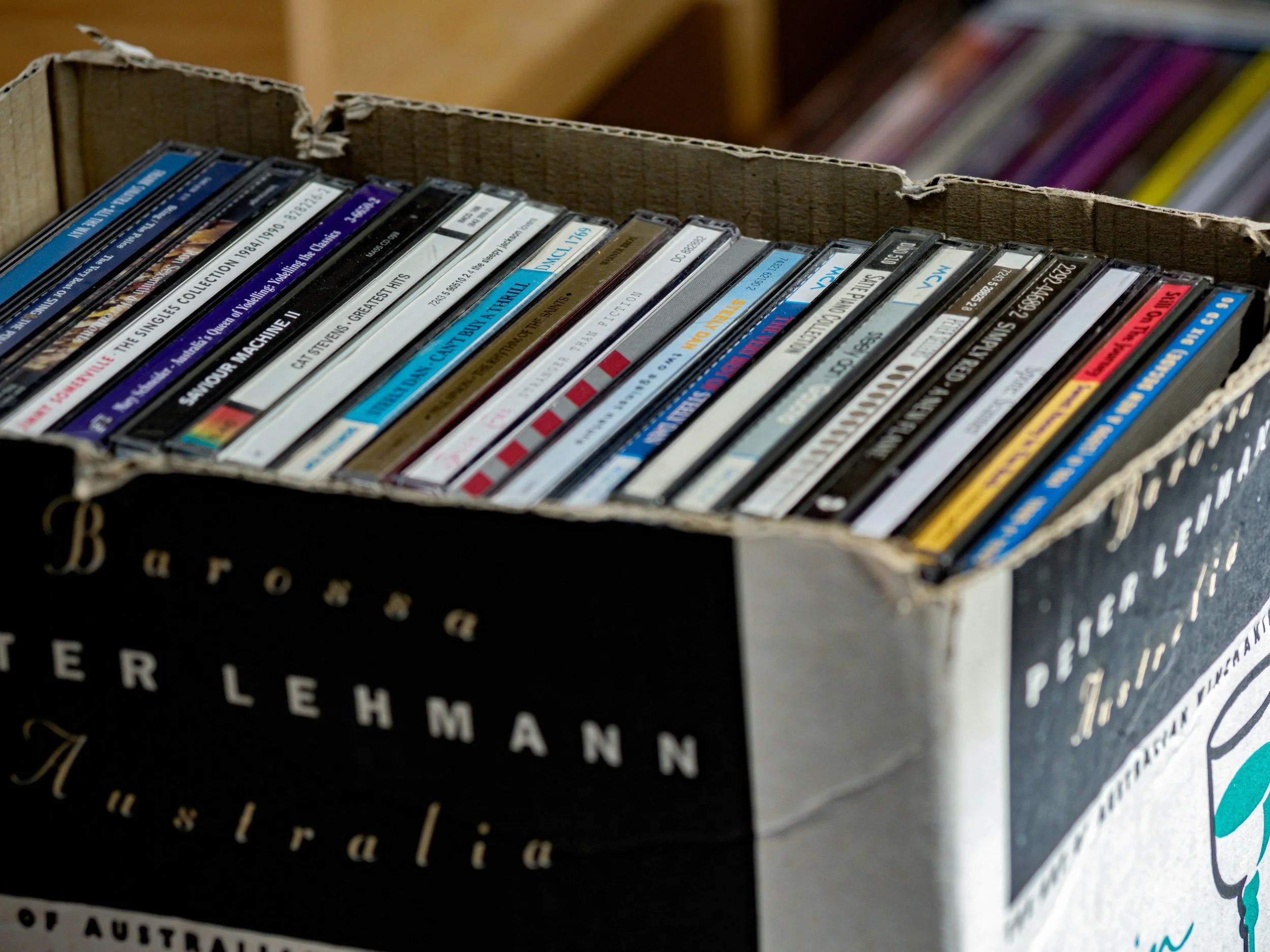 A cardboard box filled with various CDs and DVDs, with some stacked and overlapping, and a blurred background of colorful books or magazines.
