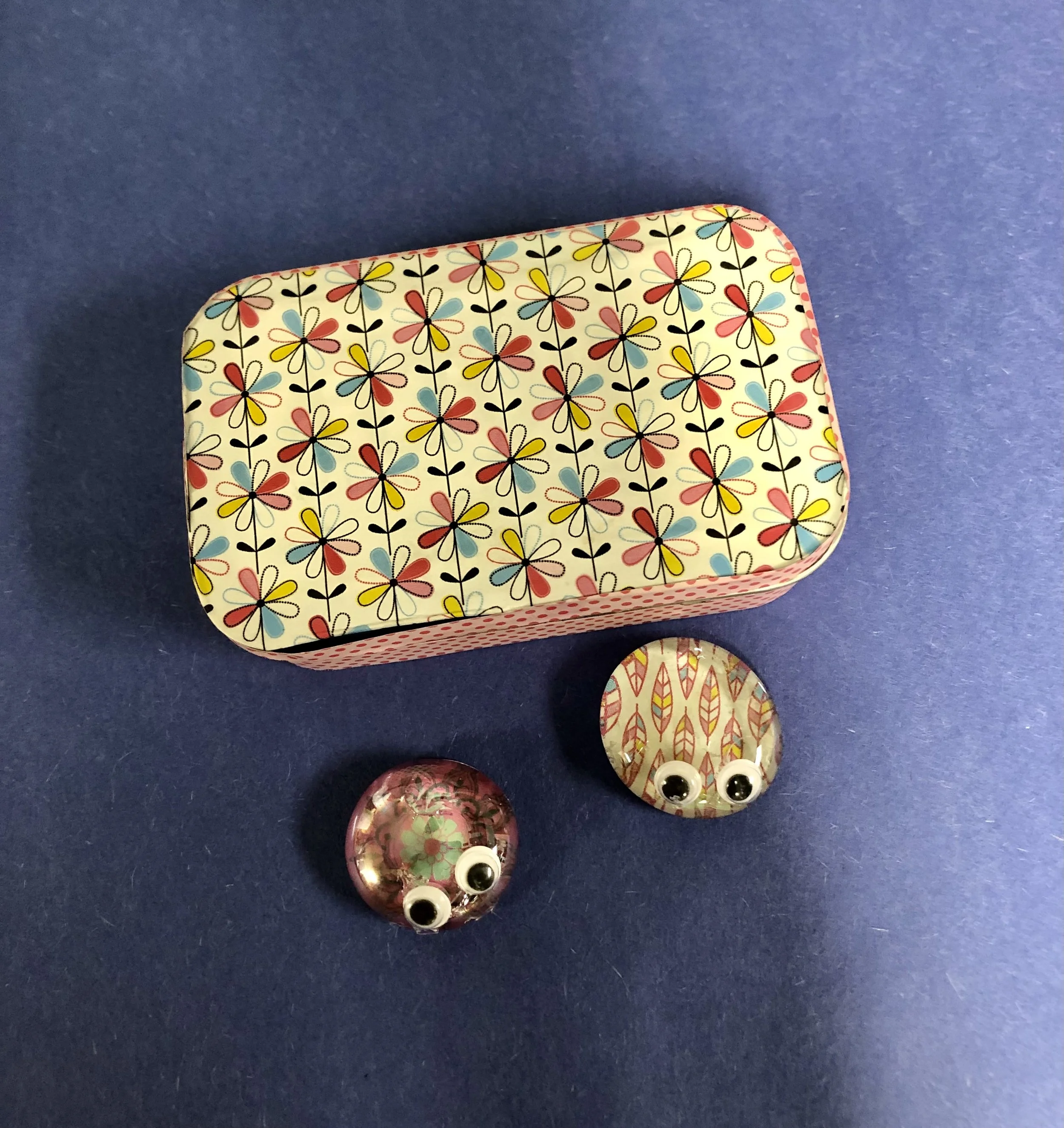 A collection of colorful buttons and paper crafts on a blue surface, including a decorated rectangular box with a floral pattern and googly eyes on a round paper craft.