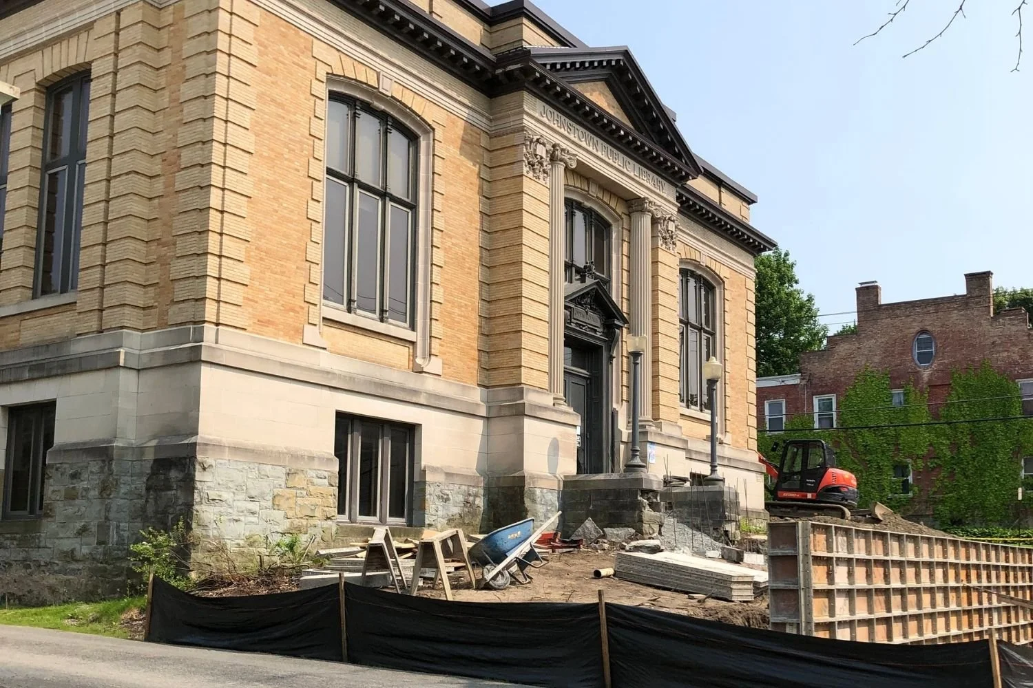 Construction site in front of a historic building labeled 'JOHNSTOWN PUBLIC LIBRARY,' with construction tools, a wheelbarrow, and a small excavator.