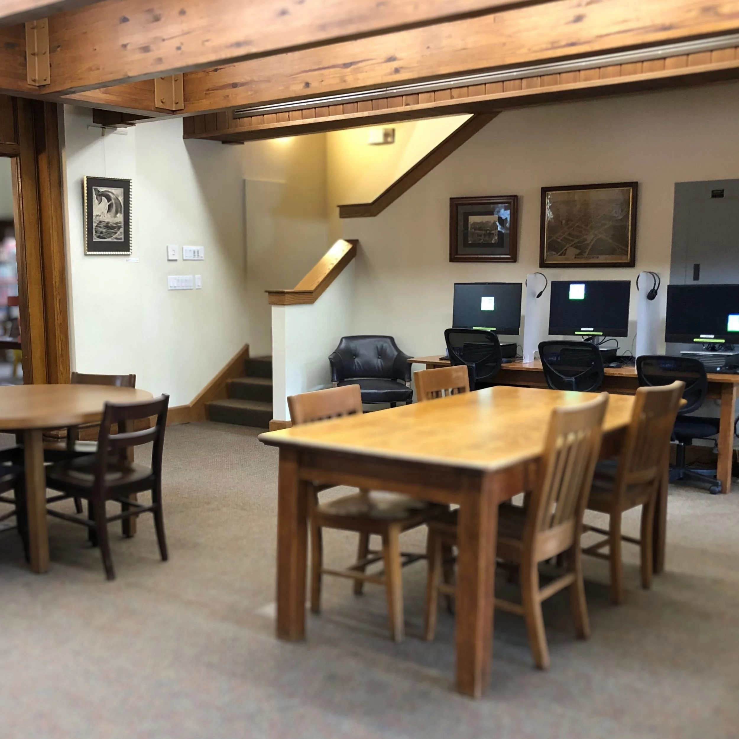 A view of a library with wooden tables and chairs, computers, and a staircase. A large leather armchair is visible on the left.