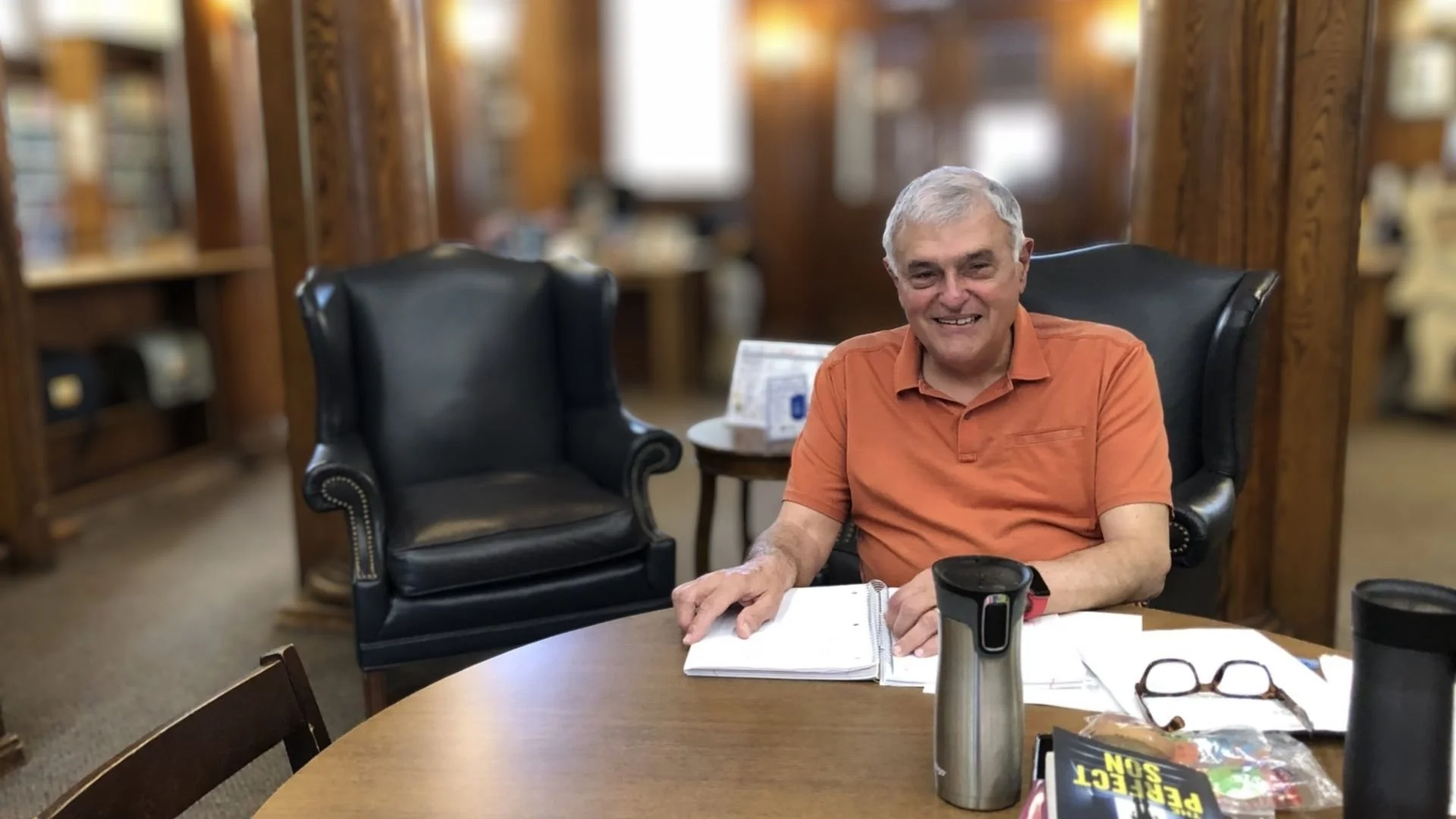 A middle-aged man with gray hair wearing an orange polo shirt sitting at a table in a library, smiling, with books and a notebook in front of him. There are bookshelves in the background.