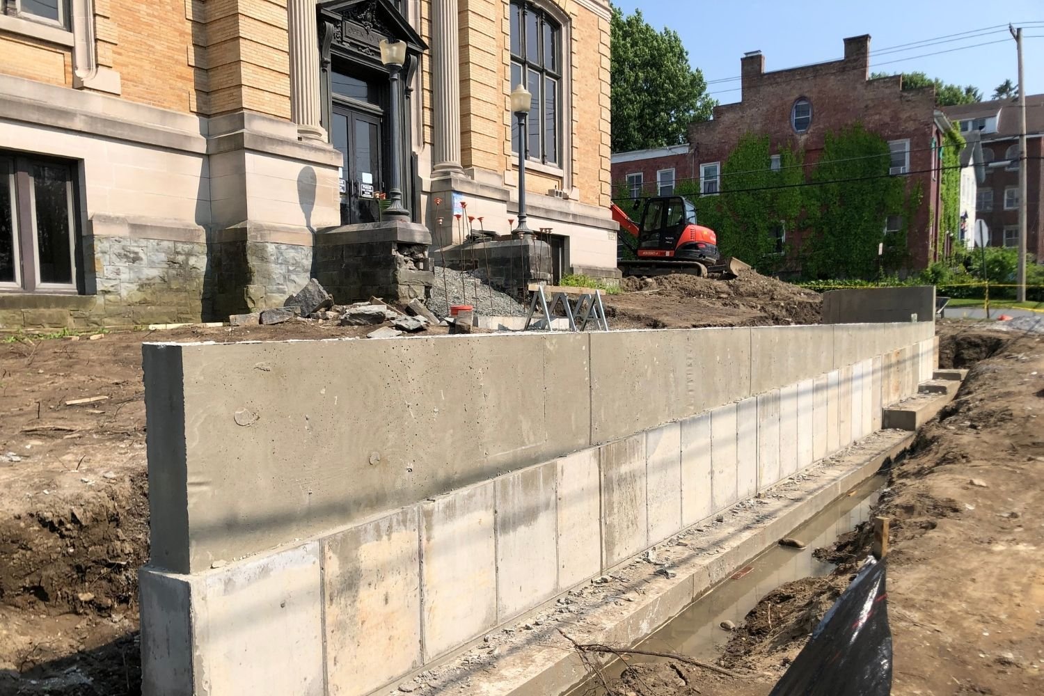 Construction site with a partially built concrete wall in front of an old building with steps and large windows, a small excavator on dirt, and a street with buildings and trees in the background.