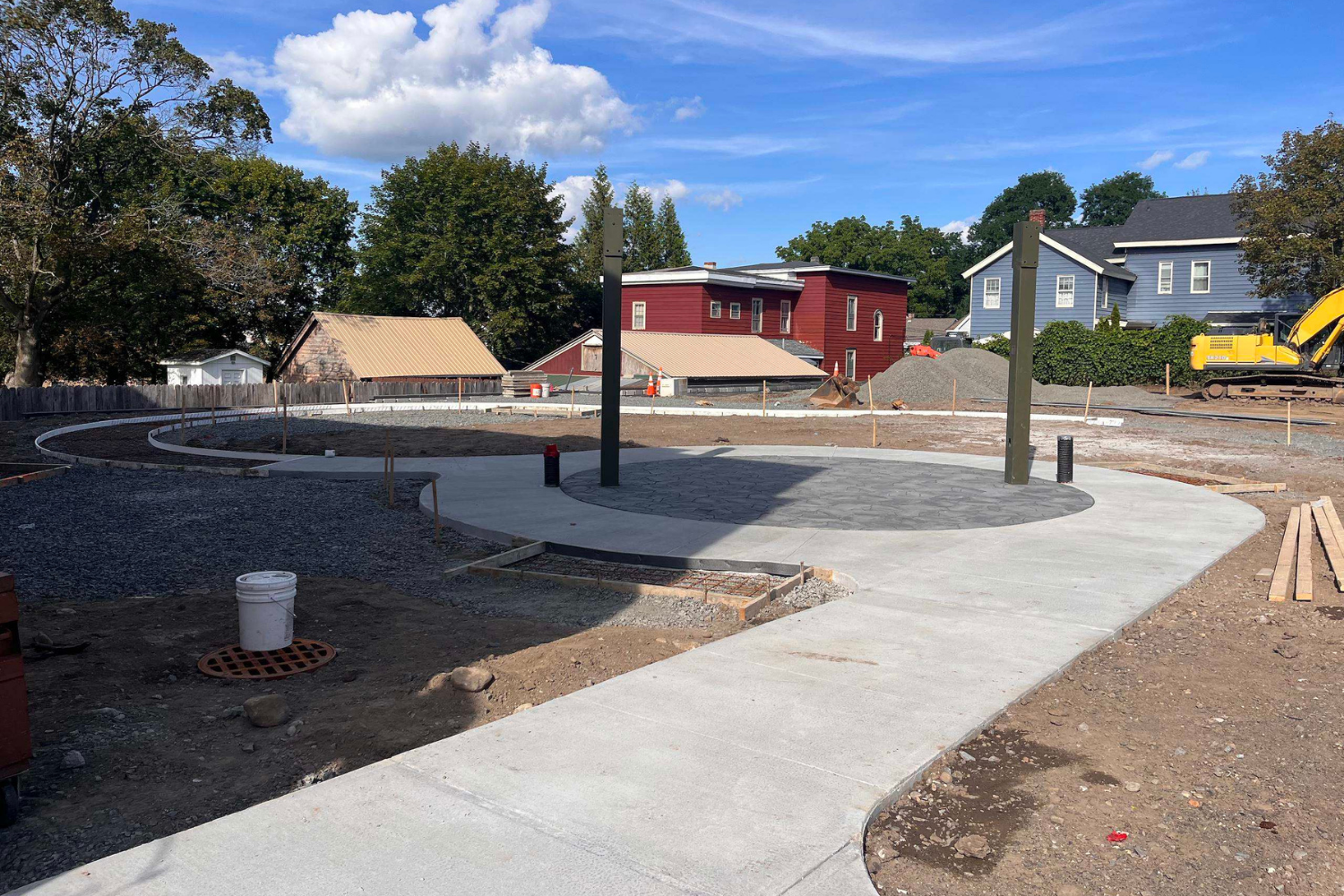 Newly constructed circular concrete pathway surrounding a large black circular pad with two posts, with other construction materials and equipment around, in a residential neighborhood.