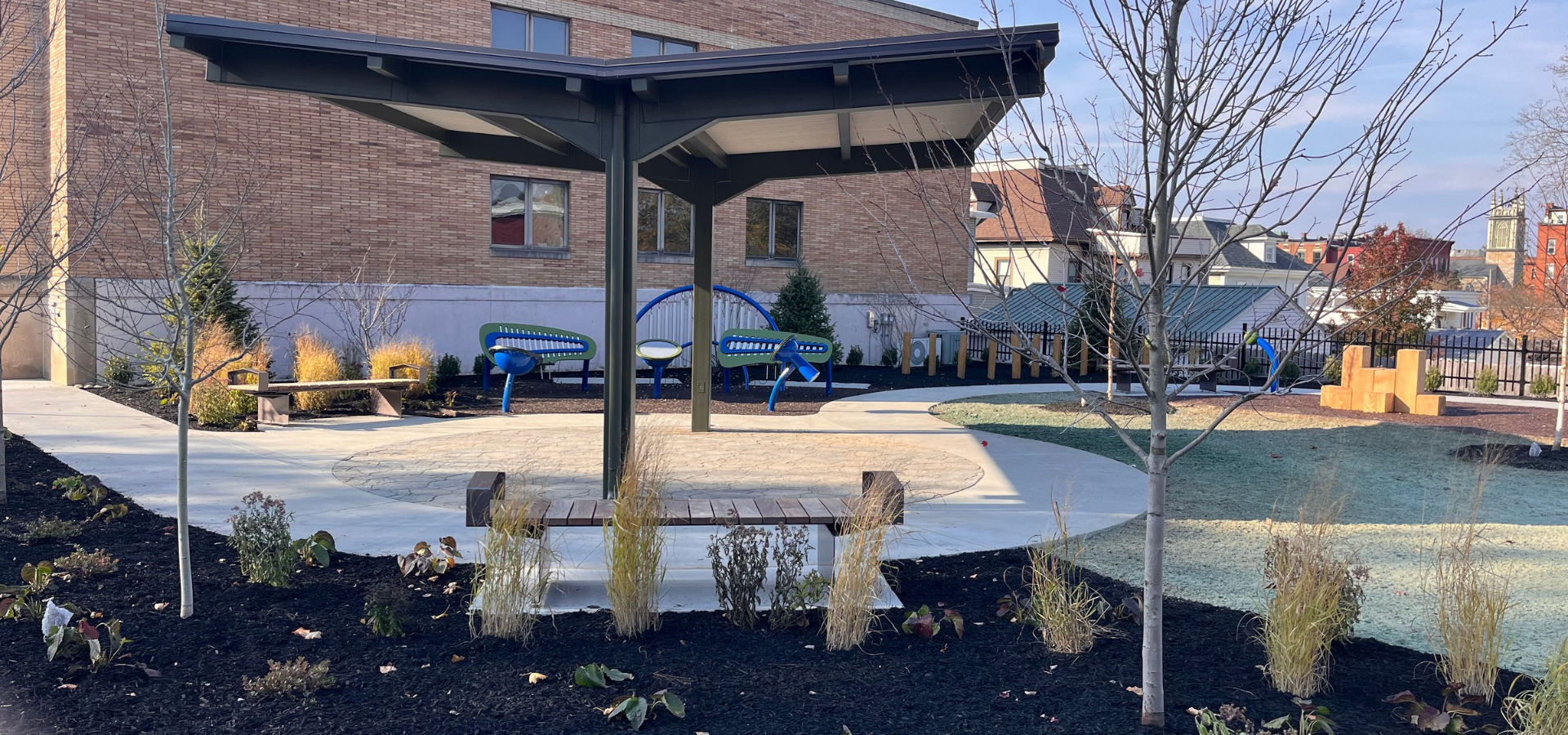 Landscaped outdoor recreation area with covered seating, benches, colorful playground equipment, paved walkways, and a brick building