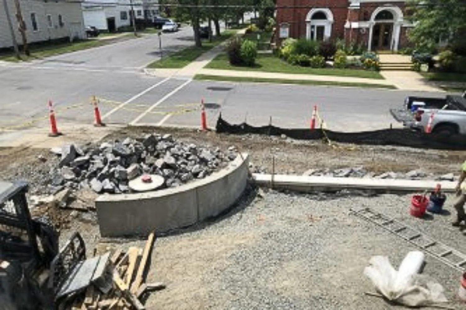 A construction site on a city street with piles of rocks, orange cones, and a partially built circular concrete structure. There is a white construction worker, and a ladder is lying on the ground nearby. The street is blocked off, and a pickup truck is parked on the side of the road.