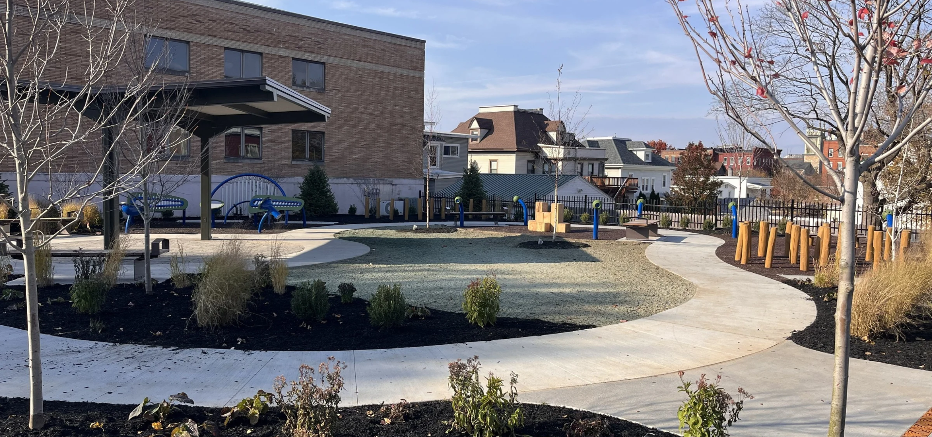 Accessible outdoor playground with blue sensory play equipment, curved concrete paths, landscaping, and young trees beside a brick building