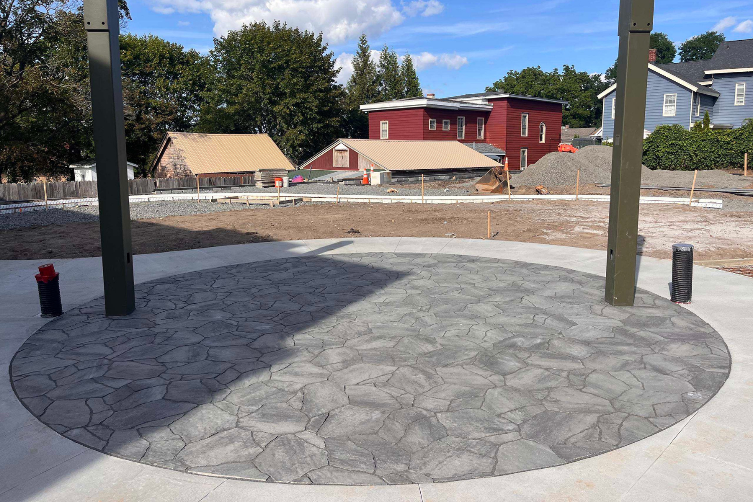 View of a construction site with a circular stamped concrete patio in the foreground and an open area of dirt behind it. In the background, there are small buildings and trees under a partly cloudy blue sky.