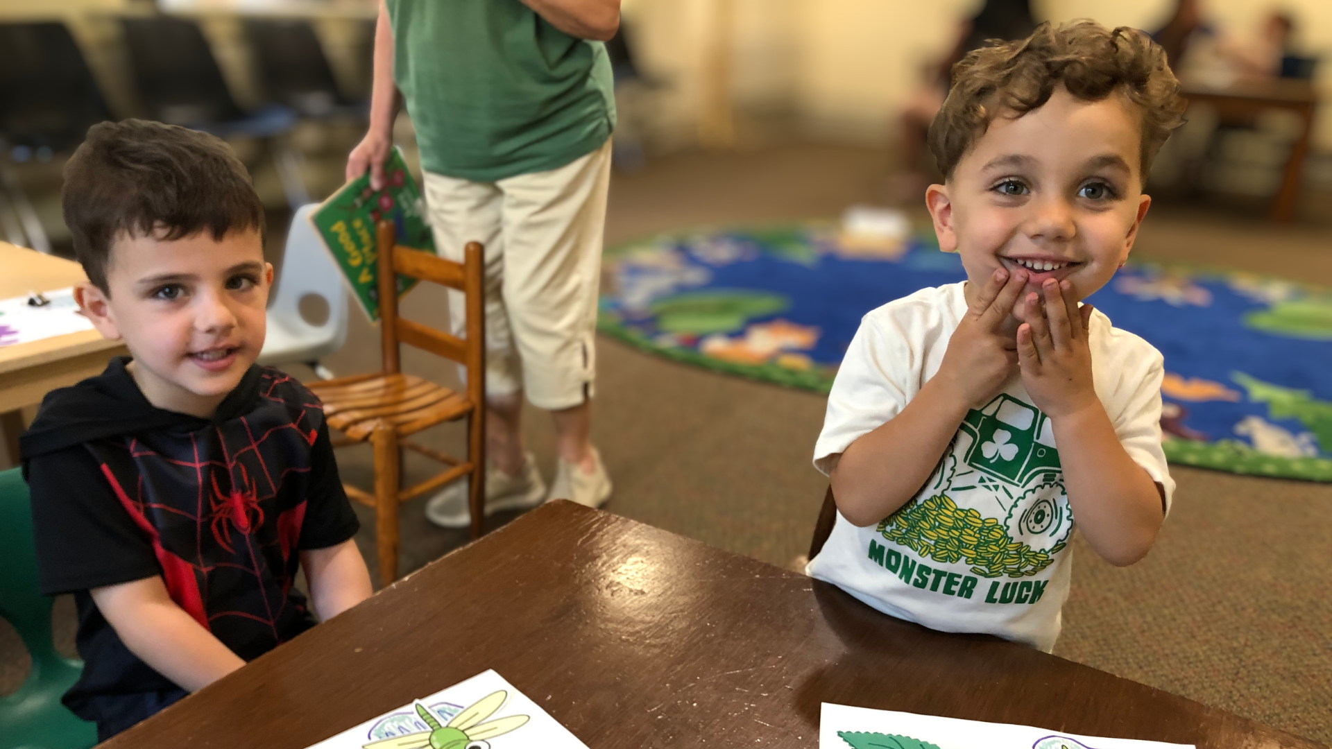 Two young boys sitting at a table in a room, one smiling and looking at the camera with hands on his face, the other looking slightly away.