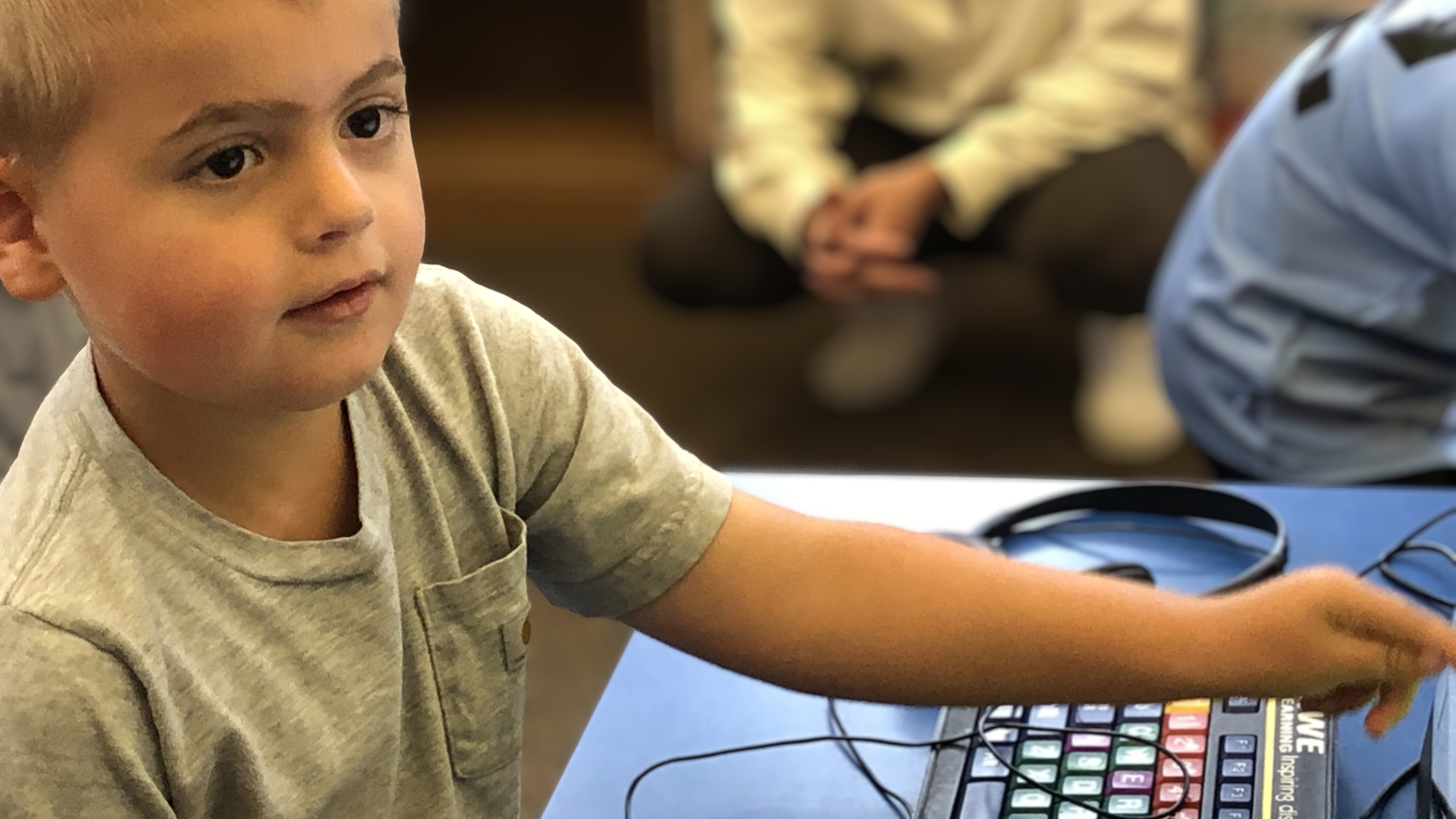 A young boy with blond hair is focusing while sitting at a table with a computer and headphones, using a keypad. There are other children in the background, some sitting on the floor.
