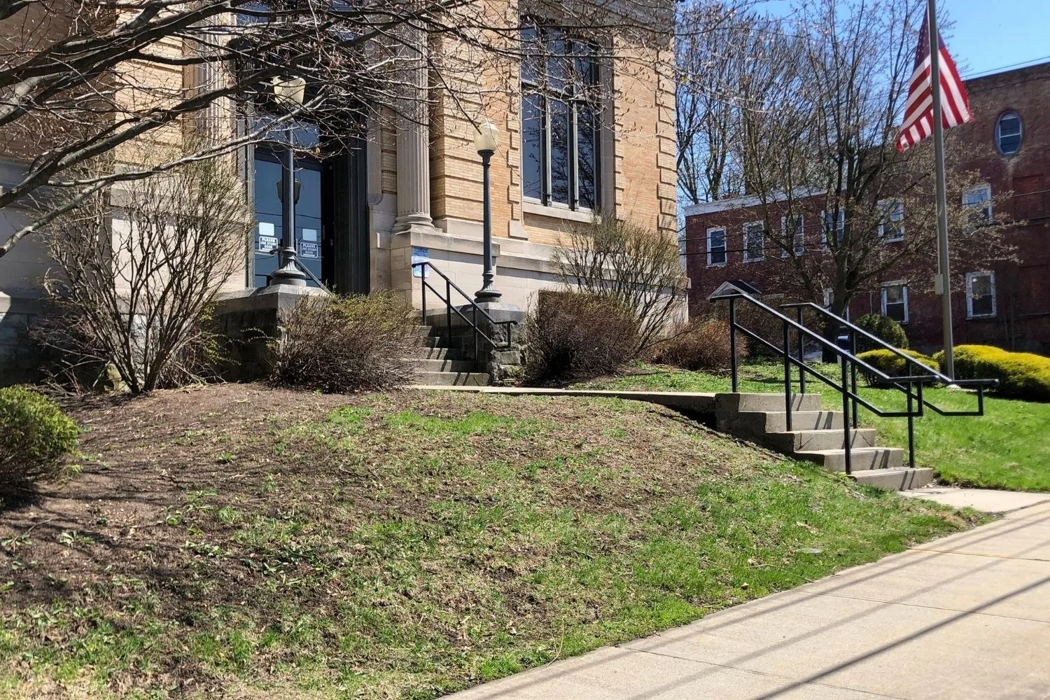 Concrete stairs with black railings leading up to a beige brick building entrance, with American flag on a flagpole nearby, surrounded by small bushes and leafless trees under a clear blue sky.