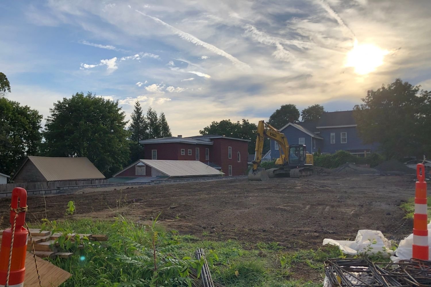 Construction site with a yellow excavator on cleared ground, surrounded by orange traffic cones and construction materials, with houses and trees in the background and a partly cloudy sky with the sun setting.