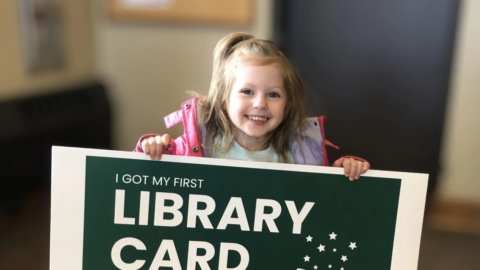A young girl with reddish hair smiles and holds a sign that reads 'I GOT MY FIRST LIBRARY CARD' at a library.