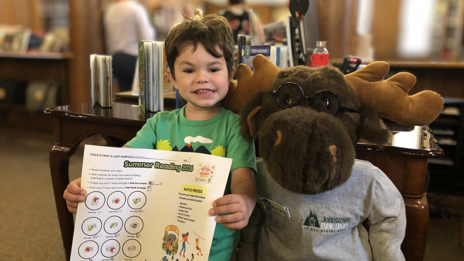 A young boy with brown hair holding a summer reading program sheet at a library. Next to him is a person wearing a moose costume with glasses. They are seated in a wooden chair with books in the background.