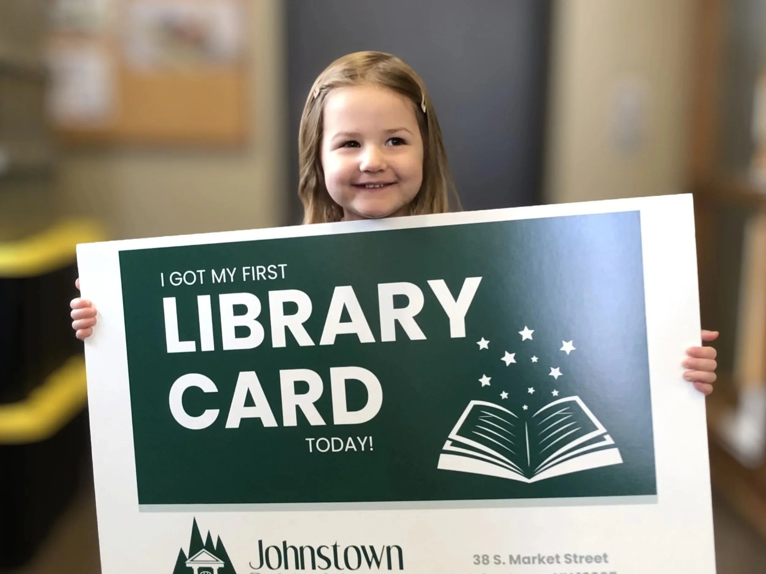 A young girl with light brown hair smiling and holding a green sign that reads, "I GOT MY FIRST LIBRARY CARD TODAY!" in a library setting.