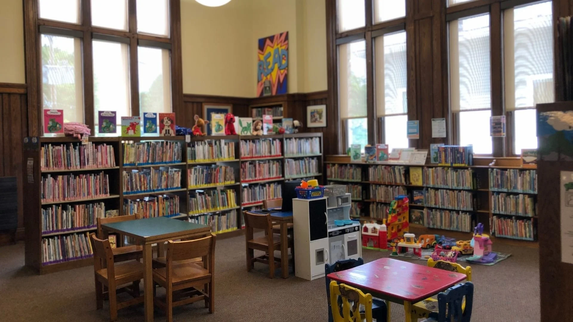 Children's section in a library with bookshelves filled with children's books, plush toys, and a play area with tables and chairs.