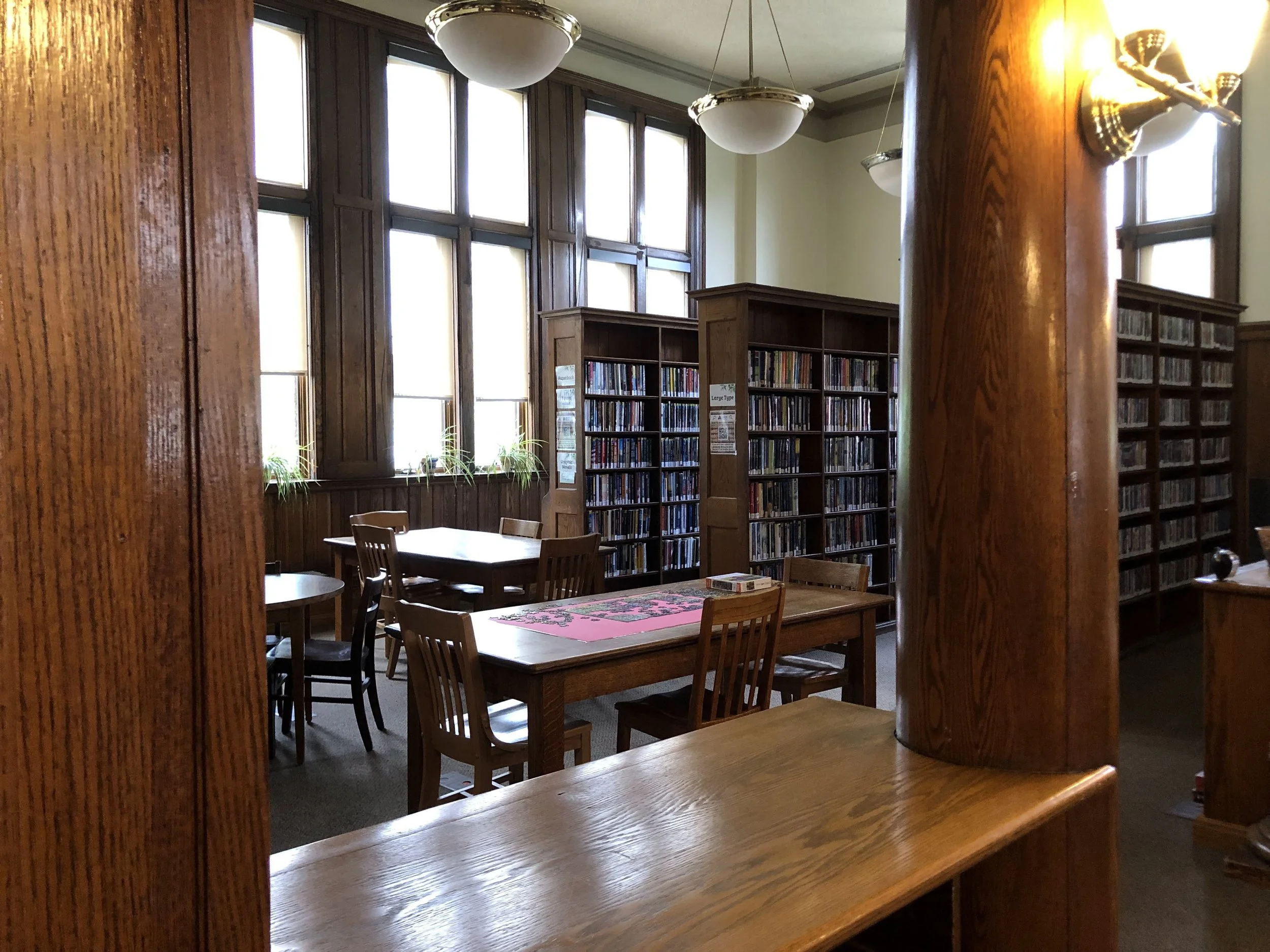 Inside a library with wooden shelves filled with books, large windows letting in natural light, and round ceiling lights. There are wooden tables and chairs for reading or studying.