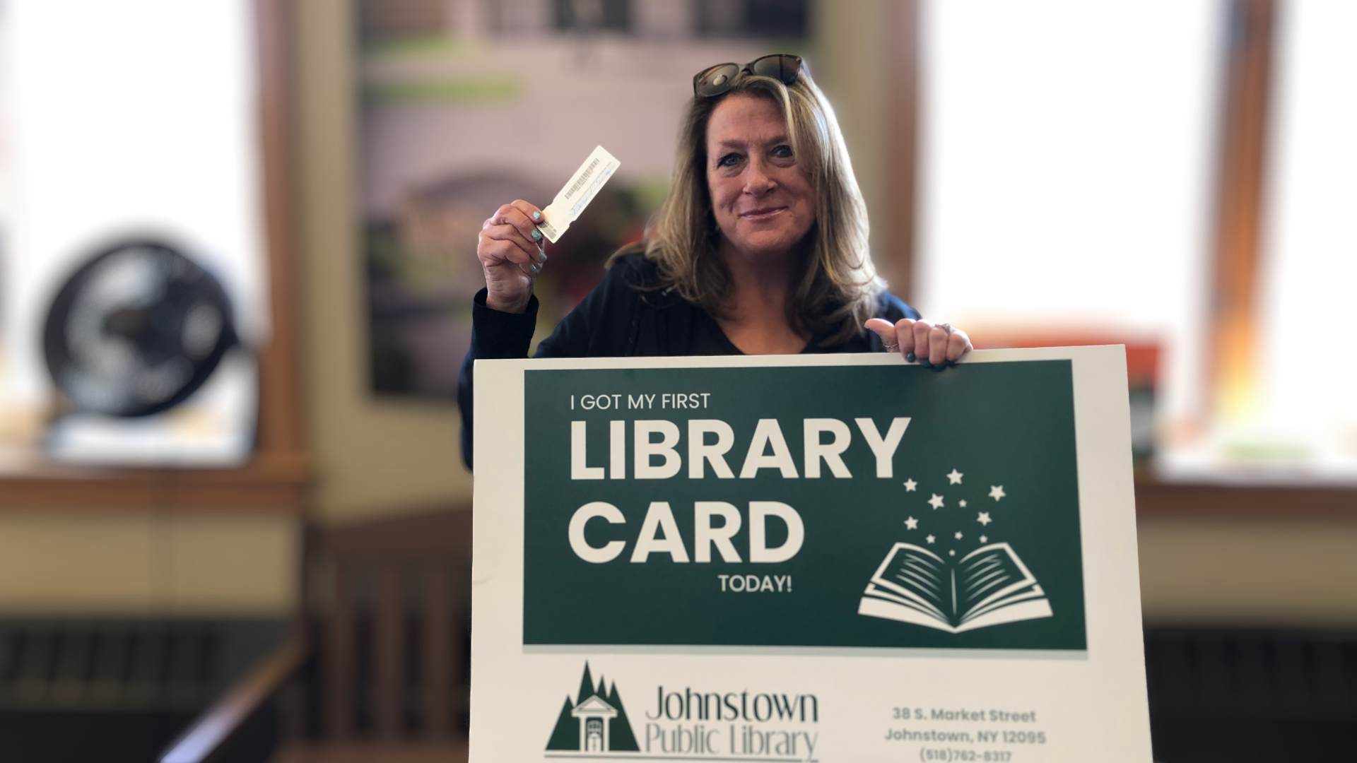 Woman holding a library card and a sign that reads 'I got my first library card today!' inside a library with a fan on a table in the background.